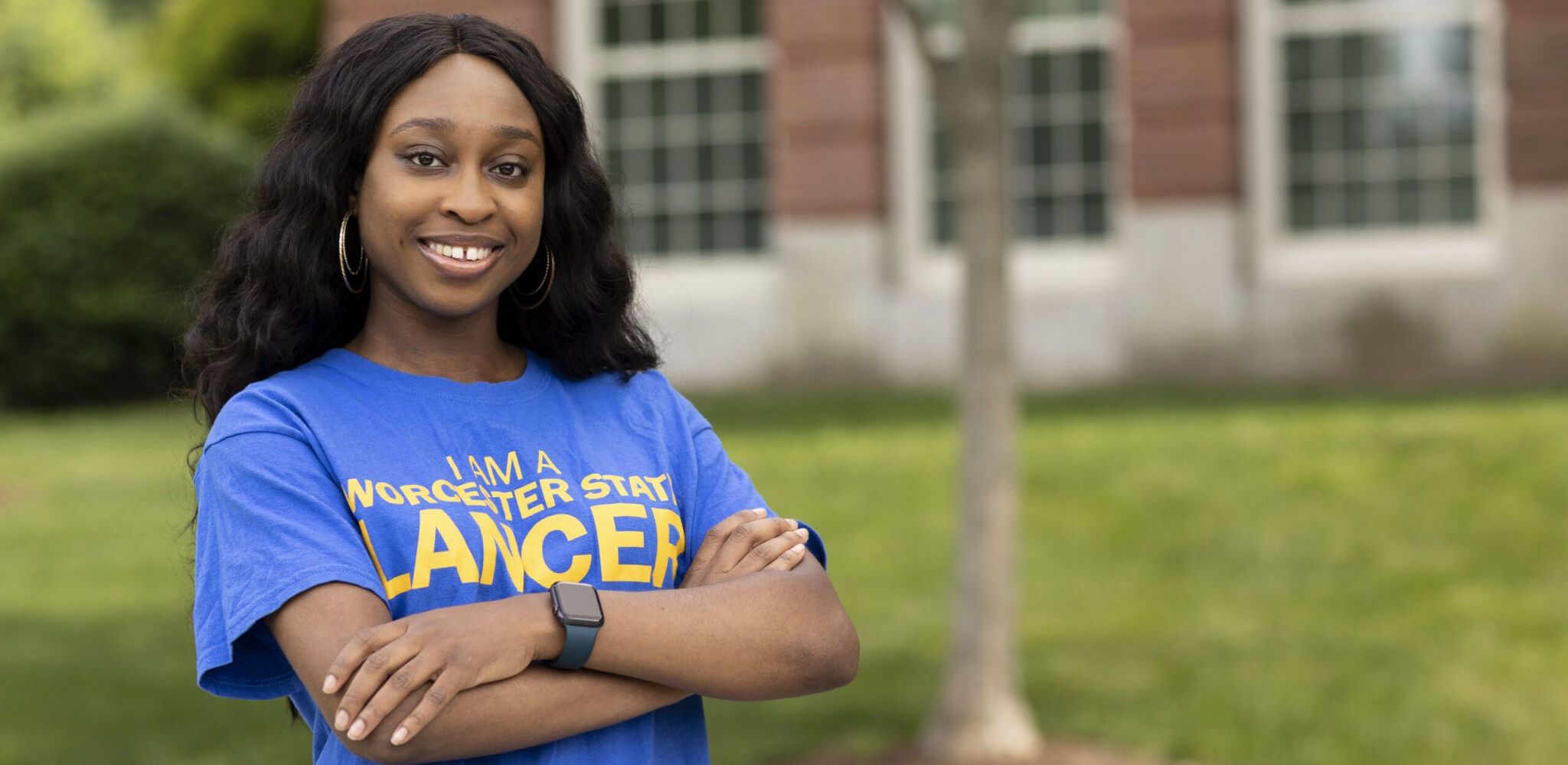 A woman stands outdoors with arms crossed, smiling, wearing a blue "Worcester State Lancer" T-shirt in front of a brick building with windows.