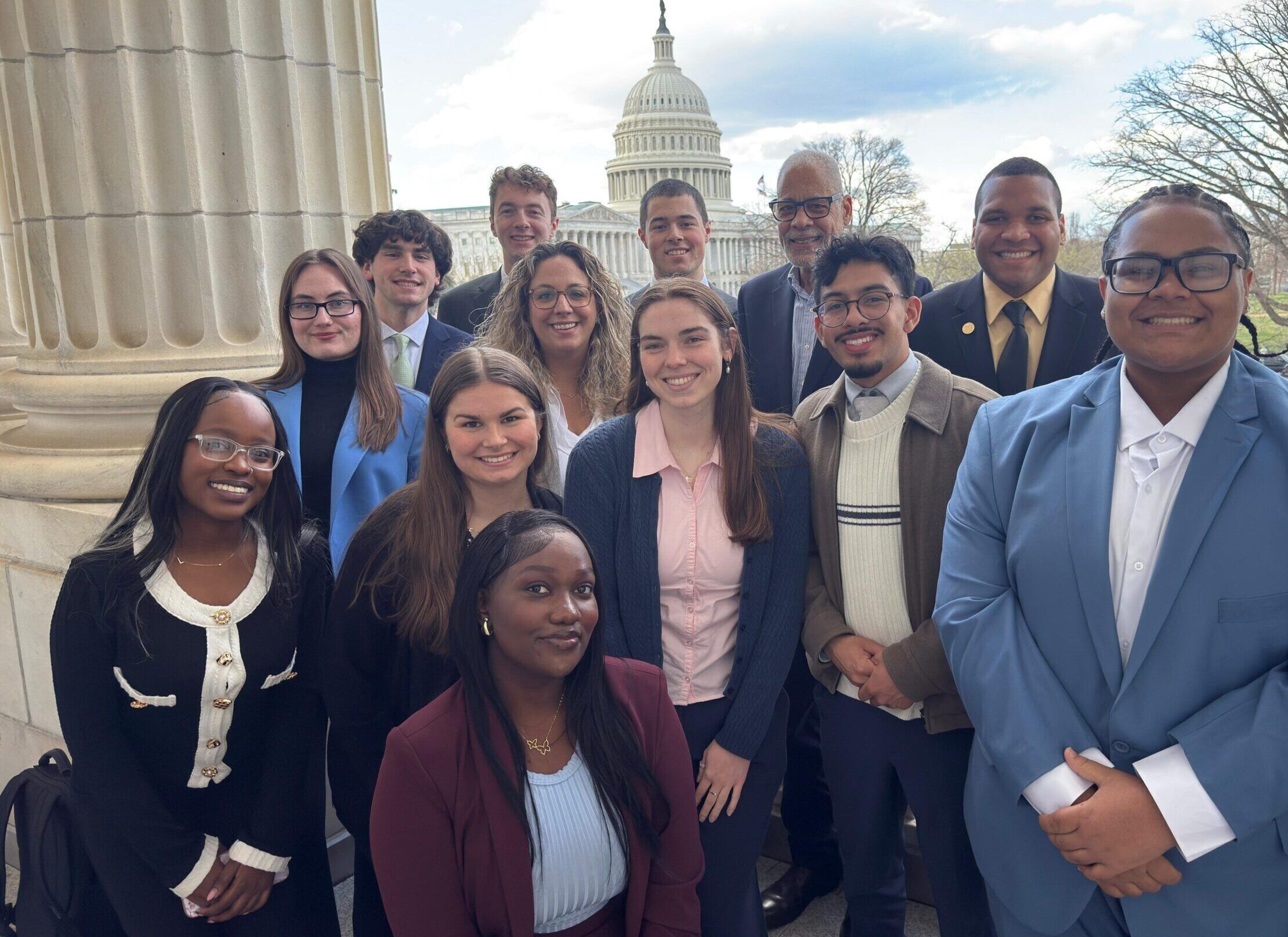 A group of thirteen people pose together on a balcony with the U.S. Capitol building and trees visible in the background.