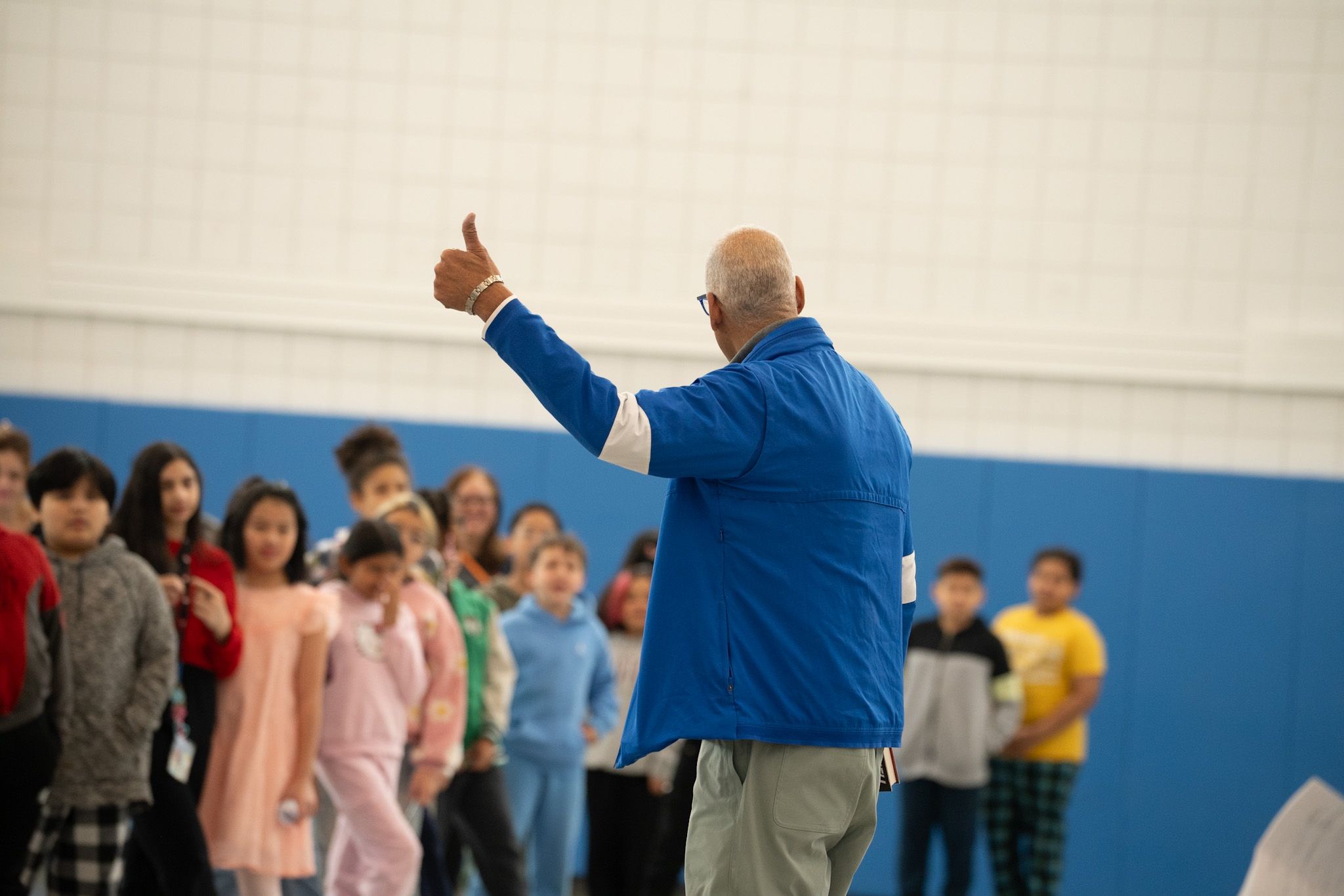 An adult in a blue jacket faces a group of children, raising a thumb in the air, inside a gymnasium with blue padded walls.