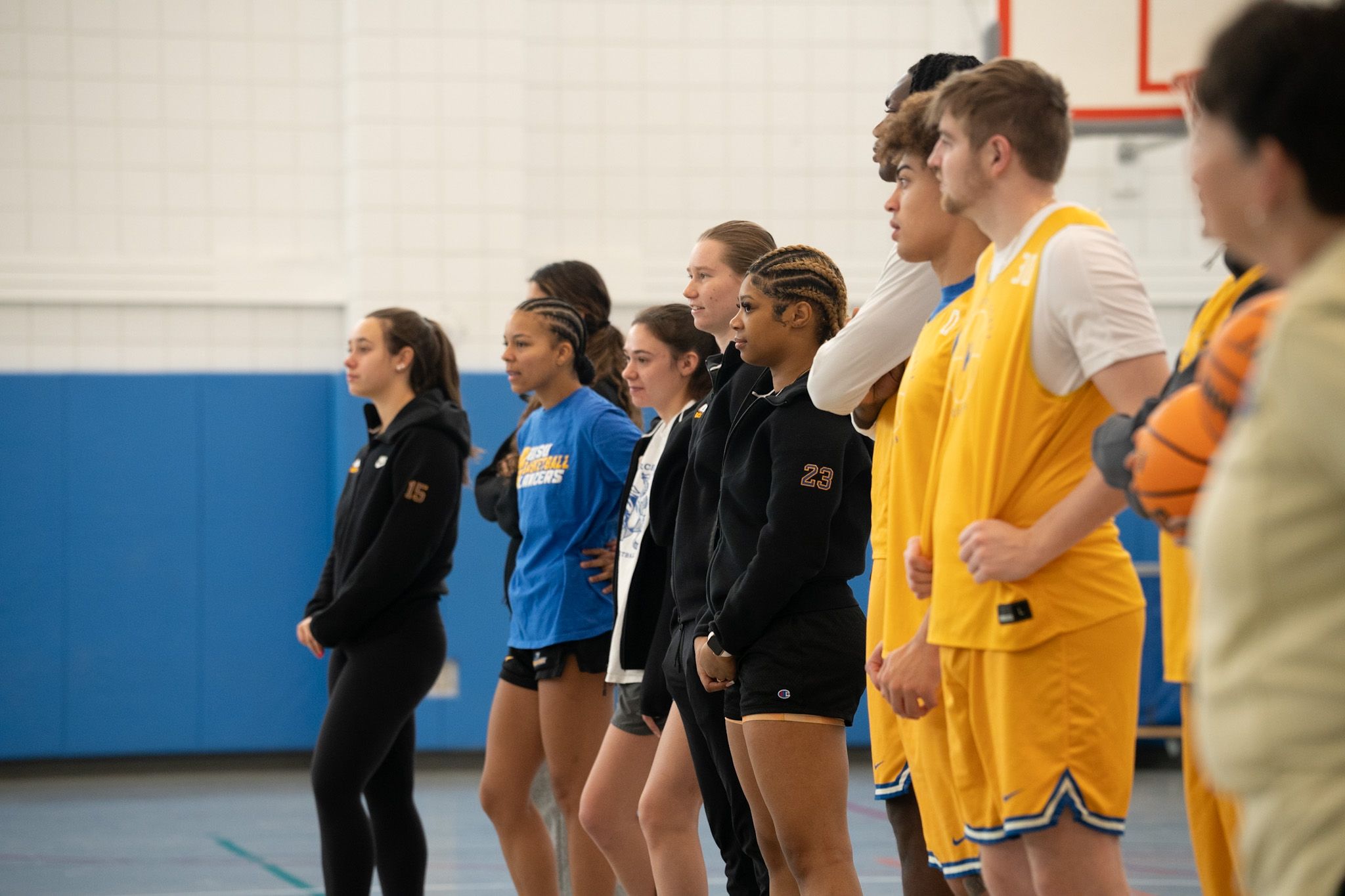 A group of basketball players, both male and female, stand in a gymnasium facing forward, wearing athletic uniforms in blue, black, and yellow.
