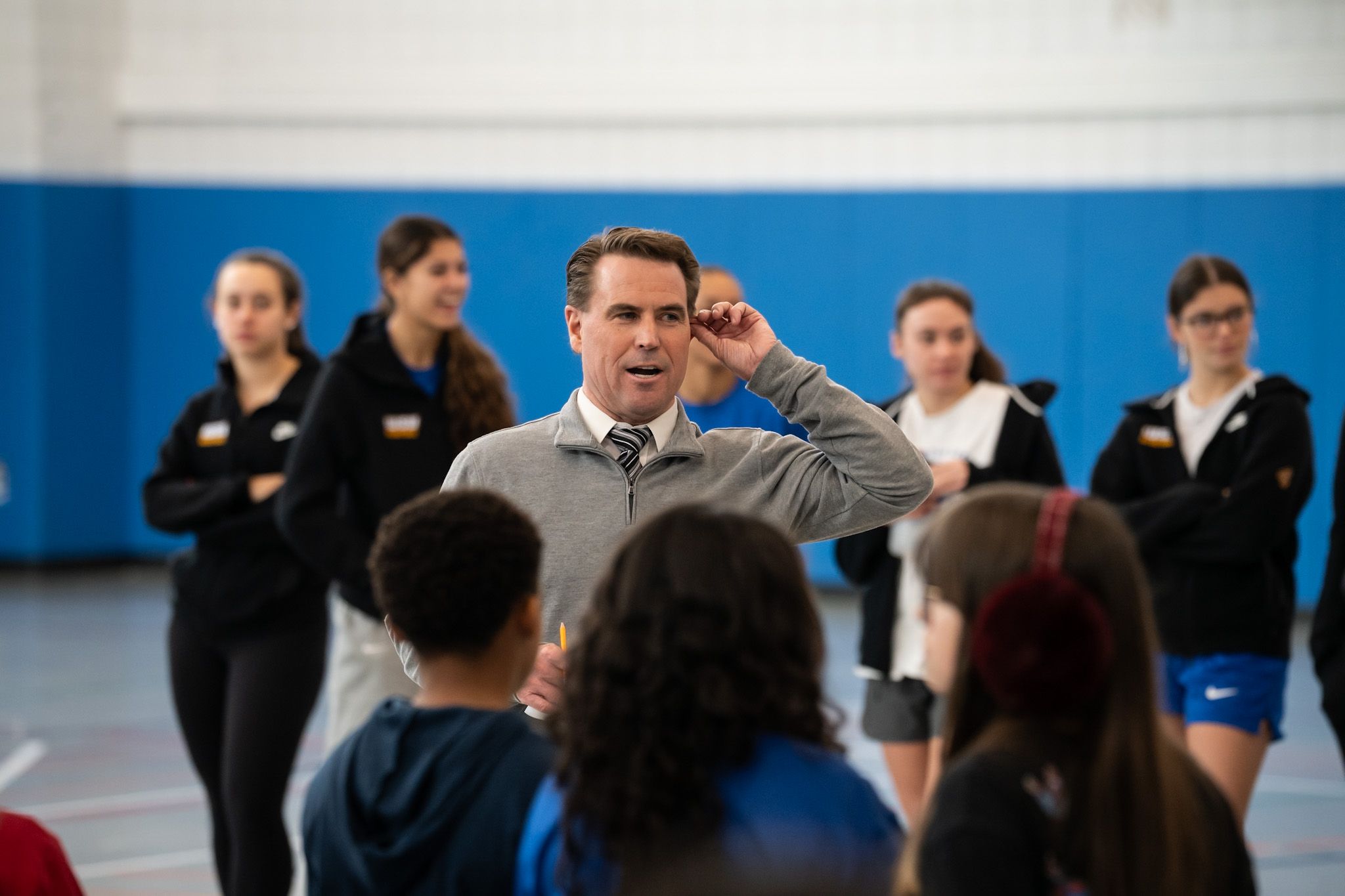 A man in a tie and sweater speaks to a group of children in a gymnasium while several women stand in the background.