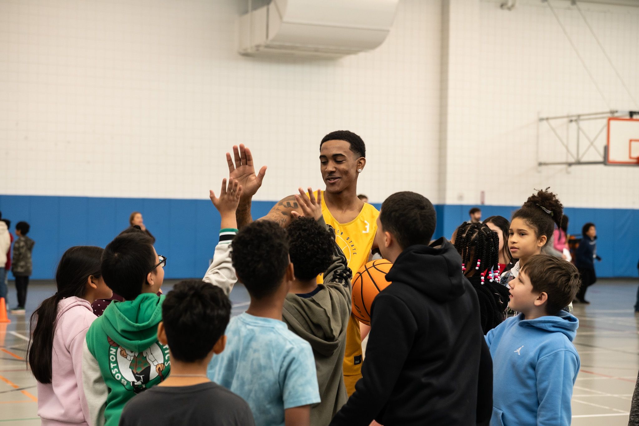 A basketball coach in a yellow jersey gives high fives to a group of children gathered around him in a gymnasium.