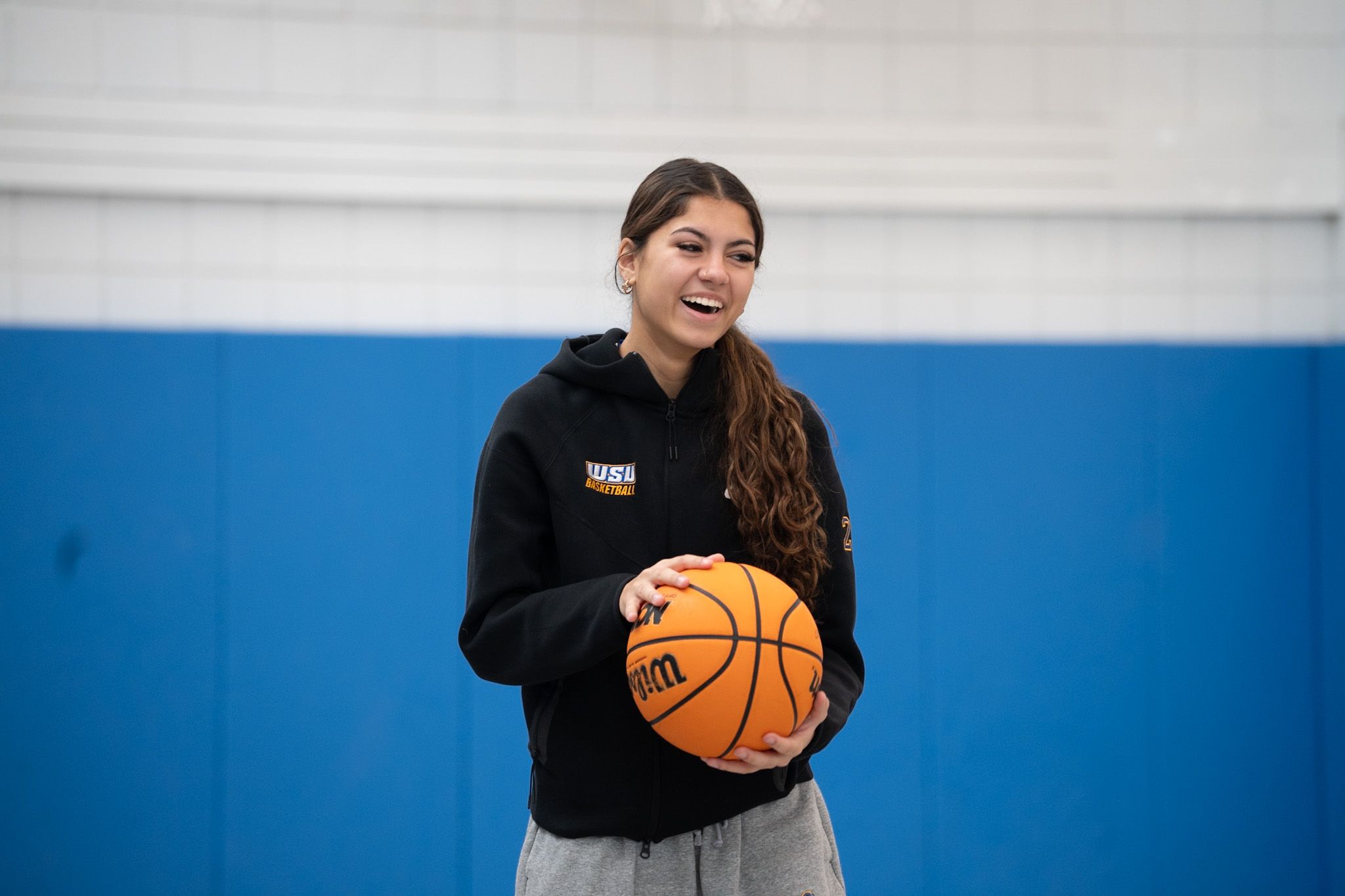 Young woman in a black hoodie holding a basketball stands in a gym with blue padded walls, smiling.