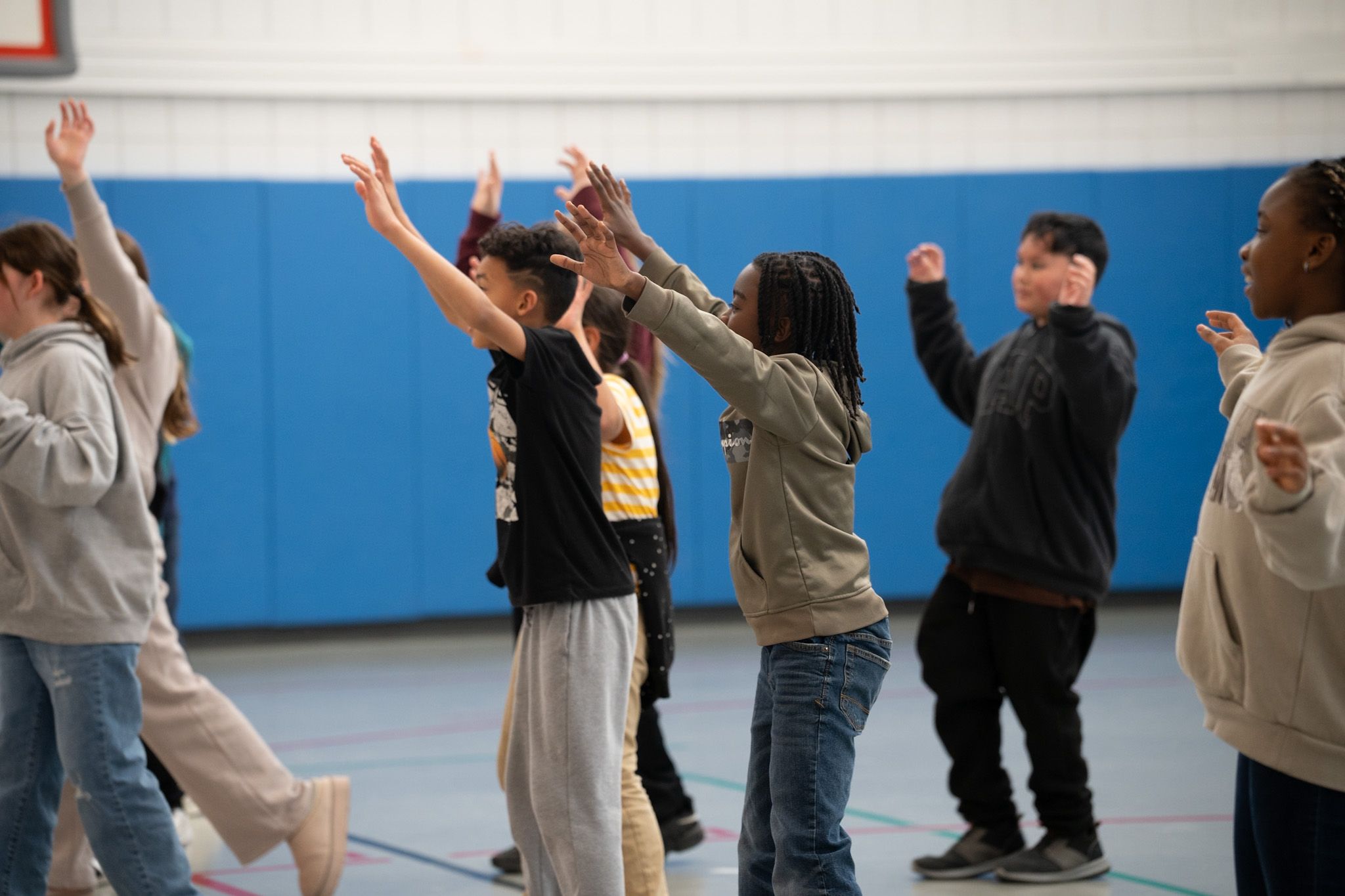 A group of children stands in a gymnasium with their arms raised, participating in what appears to be a group exercise or activity.