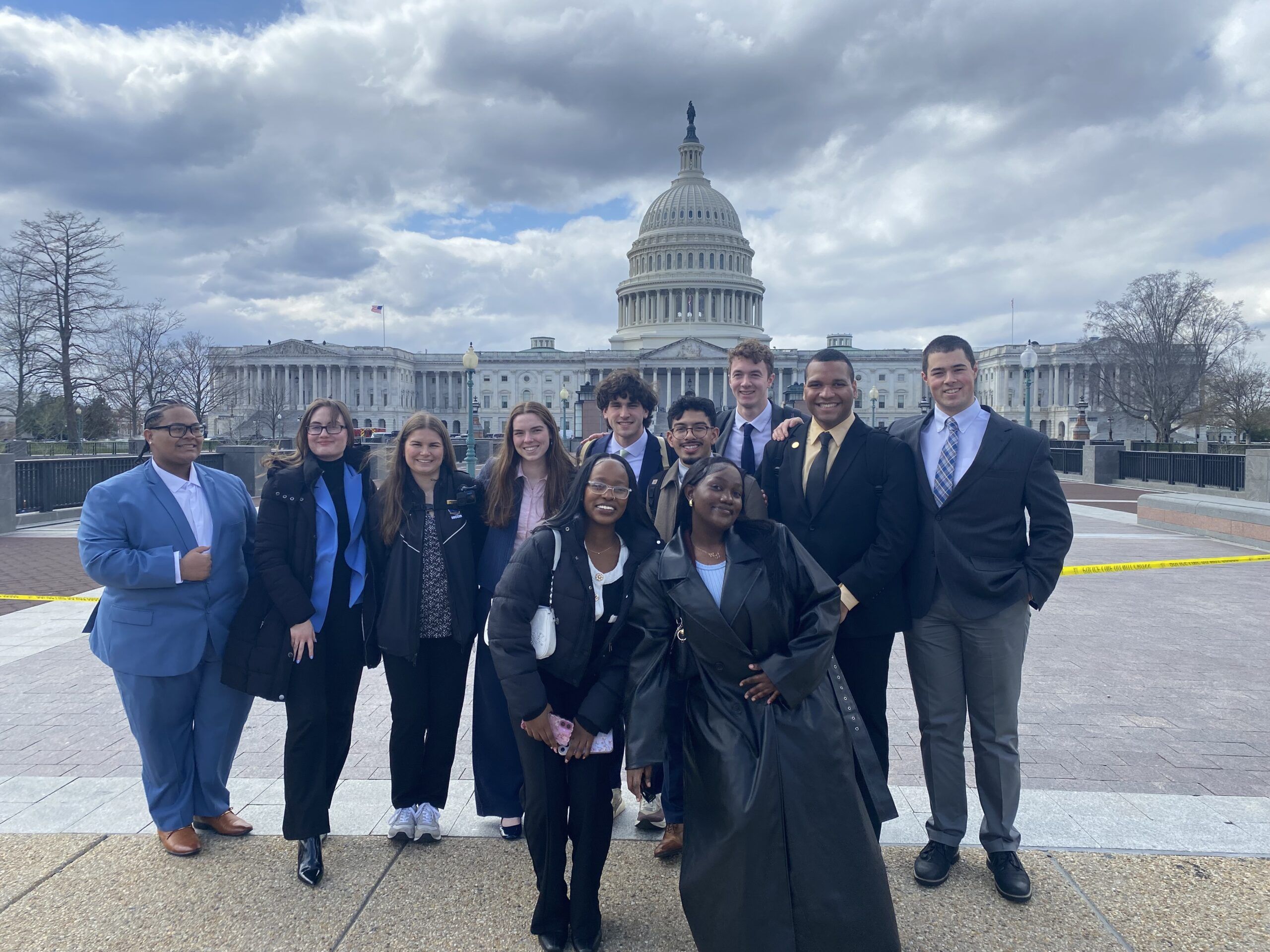 A group of ten people dressed in business attire pose and smile in front of the U.S. Capitol building on a cloudy day.