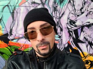 A person wearing a black beanie, sunglasses, and a black leather jacket stands in front of a colorful graffiti wall.