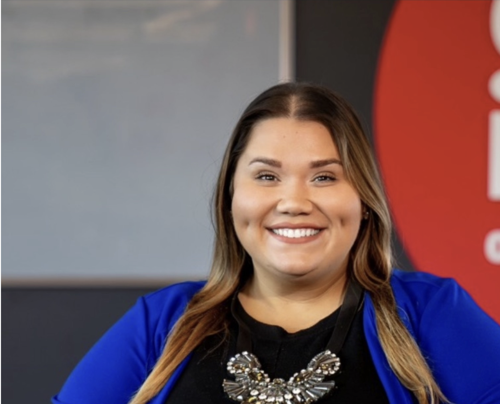 A woman with long brown hair, wearing a blue blazer and statement necklace, smiles at the camera in an office setting with a red and gray background.