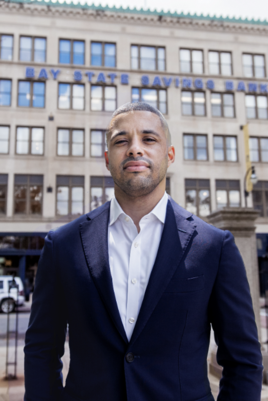 A man in a dark suit stands in front of a building with "BAY STATE SAVINGS BANK" displayed on its facade.