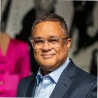 A man wearing glasses and a navy suit with a light blue shirt smiles at the camera. The background is blurred with people and a bright pink garment visible.