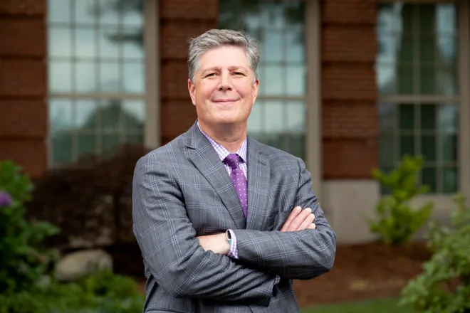 A man in a gray plaid suit and purple tie stands outdoors with arms crossed, in front of a brick building with large windows and greenery.