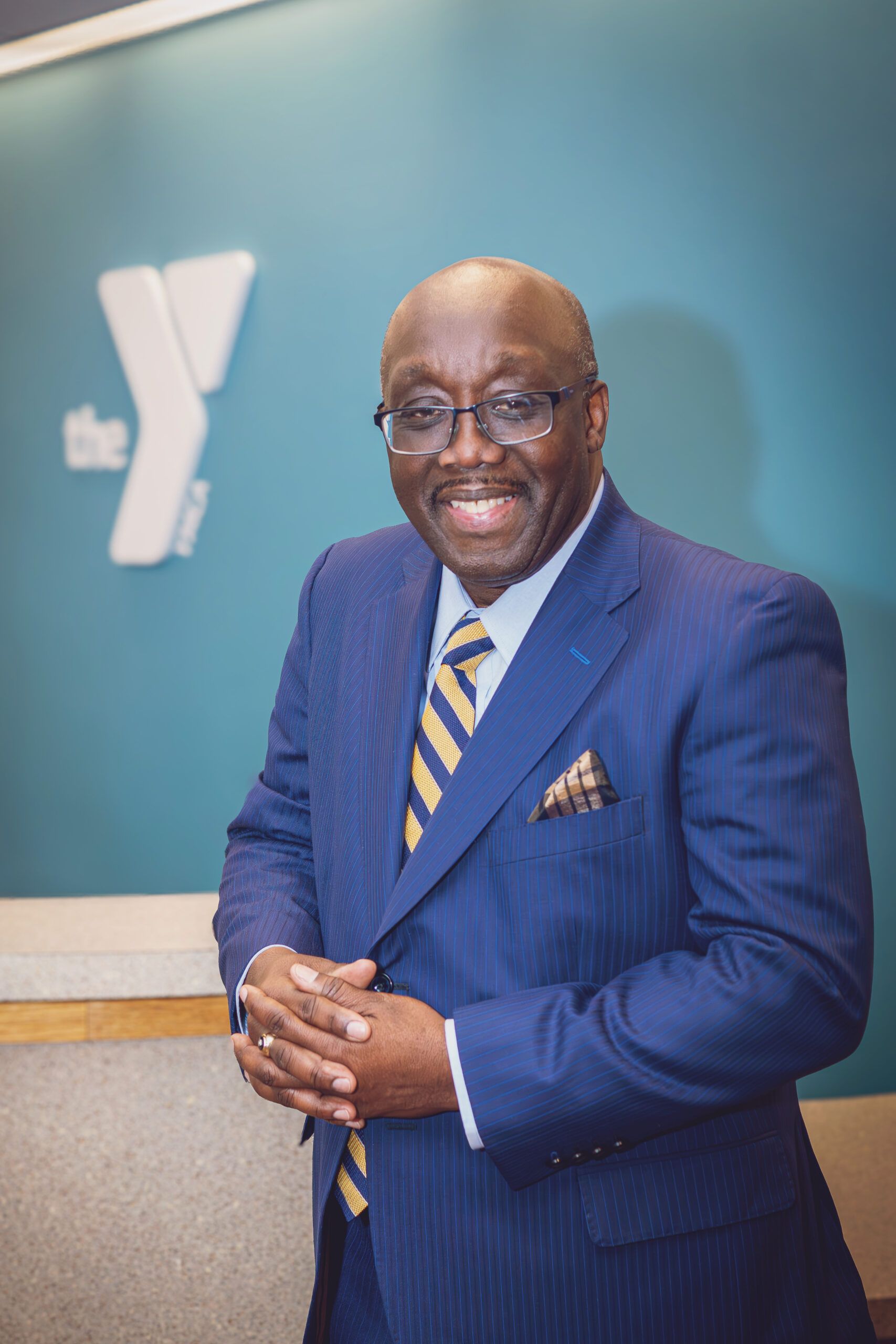 A man in a blue suit and striped tie stands and smiles in front of a desk, with a YMCA logo visible on a teal wall behind him.