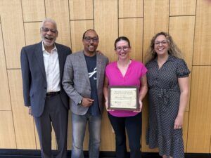 Four people stand in front of a wooden wall; the woman second from right holds a framed certificate and all are smiling at the camera.