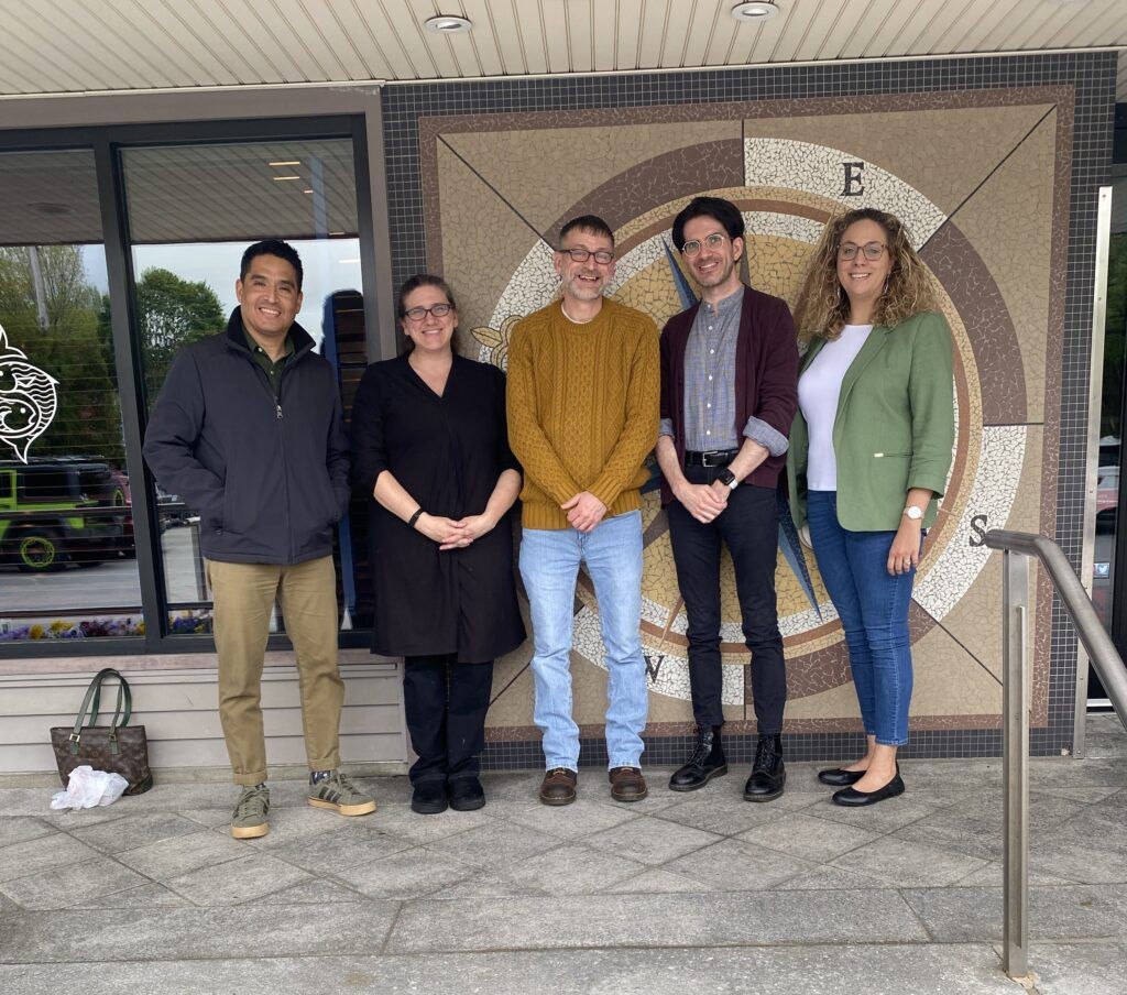 Five people stand side by side, smiling, in front of a wall with a large compass design.