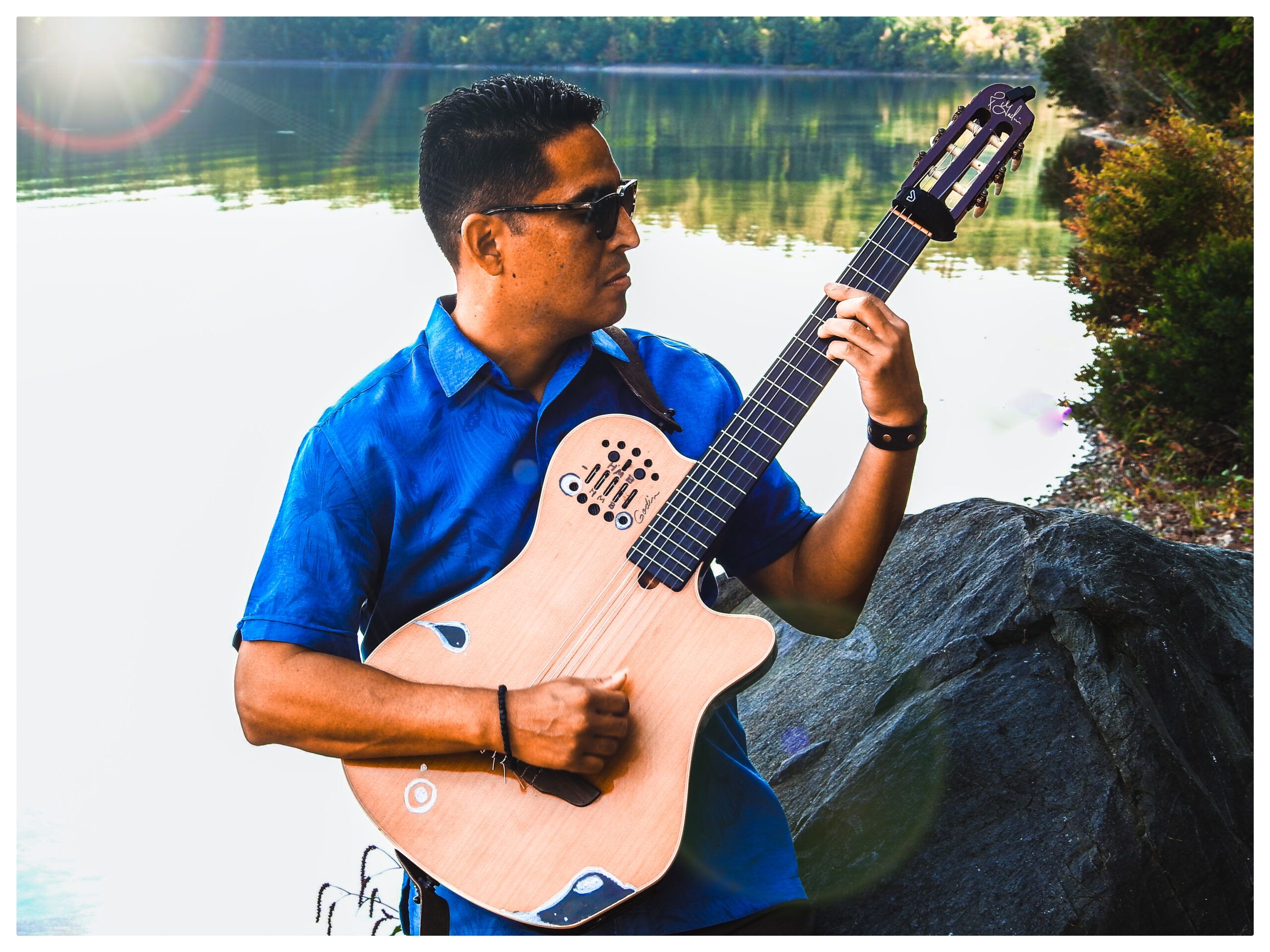 A man in a blue shirt and sunglasses plays an acoustic guitar outdoors near a lake with trees and rocks in the background.
