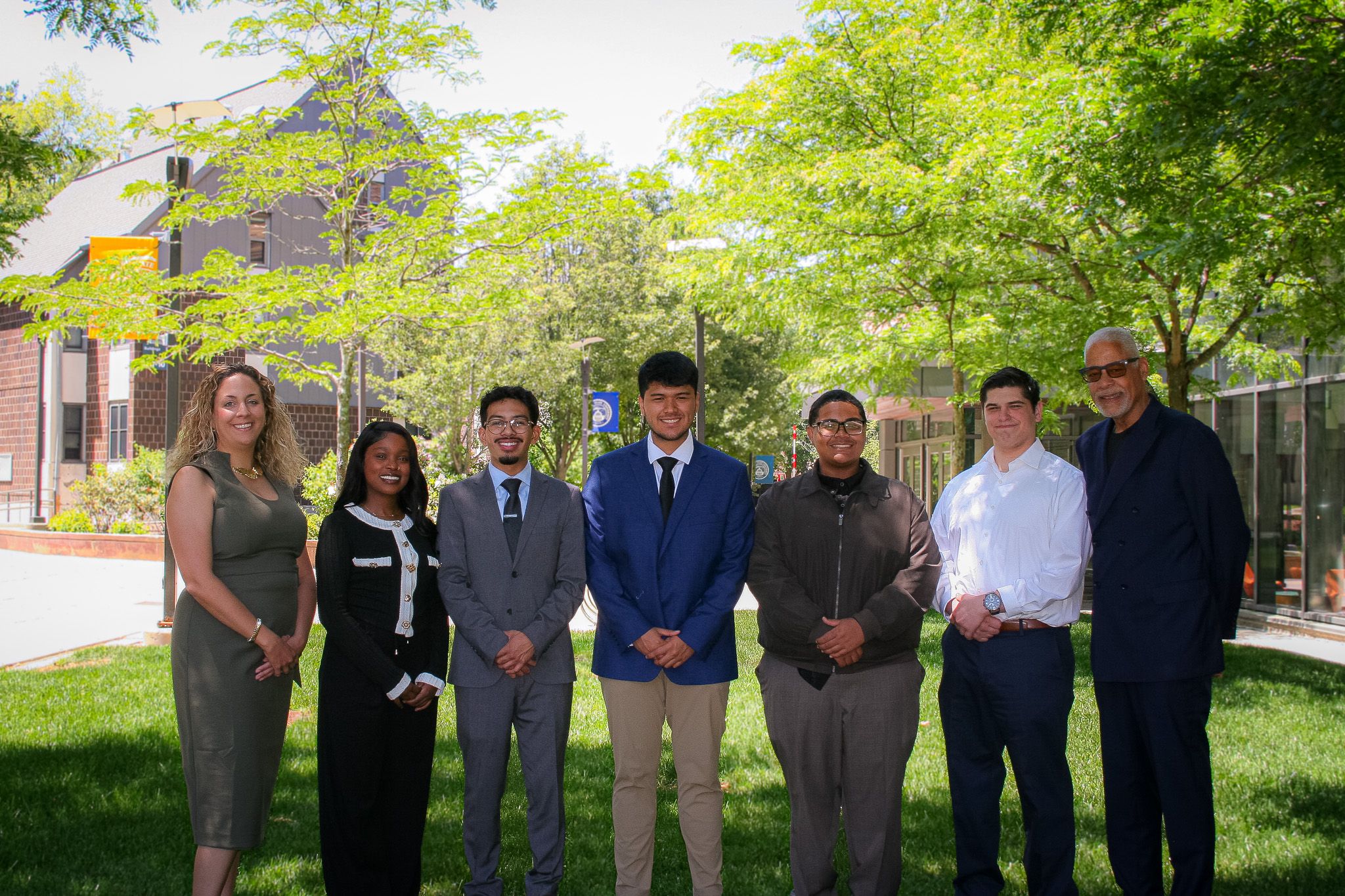 Seven people stand in a row outdoors on a sunny day, dressed in business and semi-formal attire, with trees and buildings in the background.