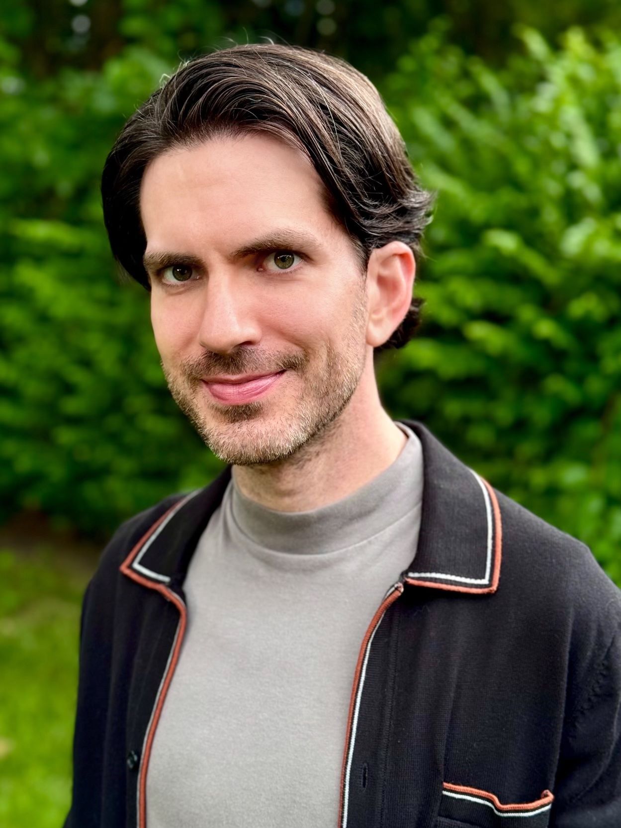 A man with dark hair and a trimmed beard is standing outdoors in front of green foliage, wearing a light gray shirt and a black jacket with white and orange trim.