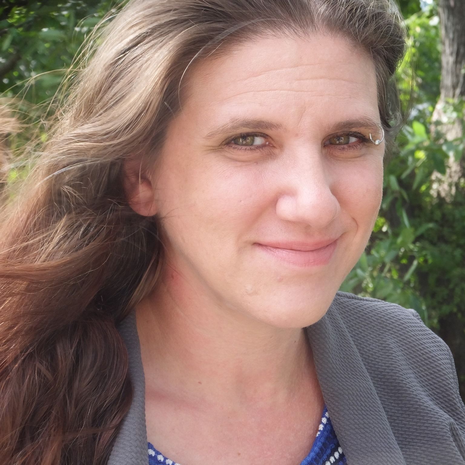 A woman with long brown hair and a gray blazer smiles slightly outdoors, with green foliage in the background.