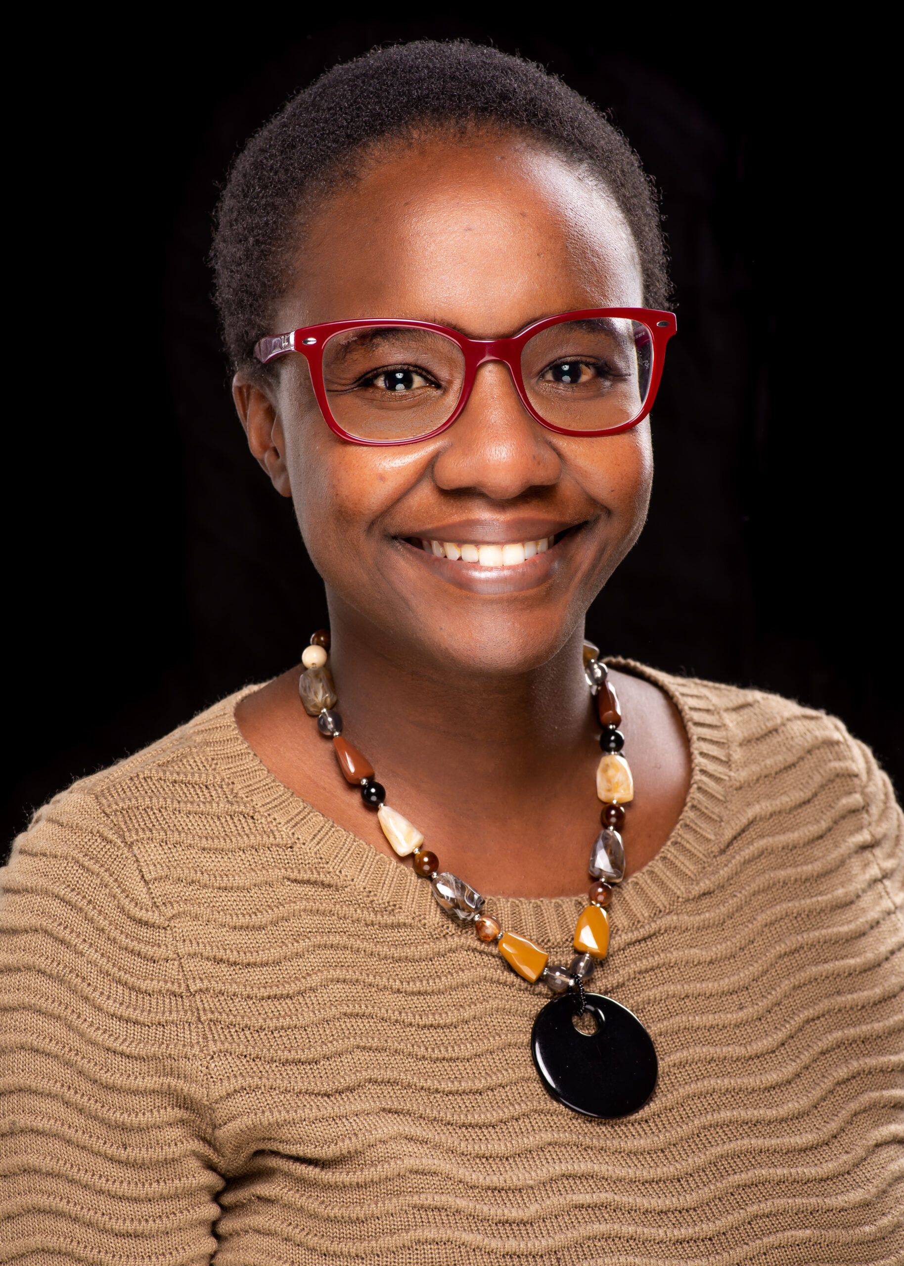 Smiling person wearing red glasses, a tan textured sweater, and a beaded necklace with a large black pendant, set against a black background.