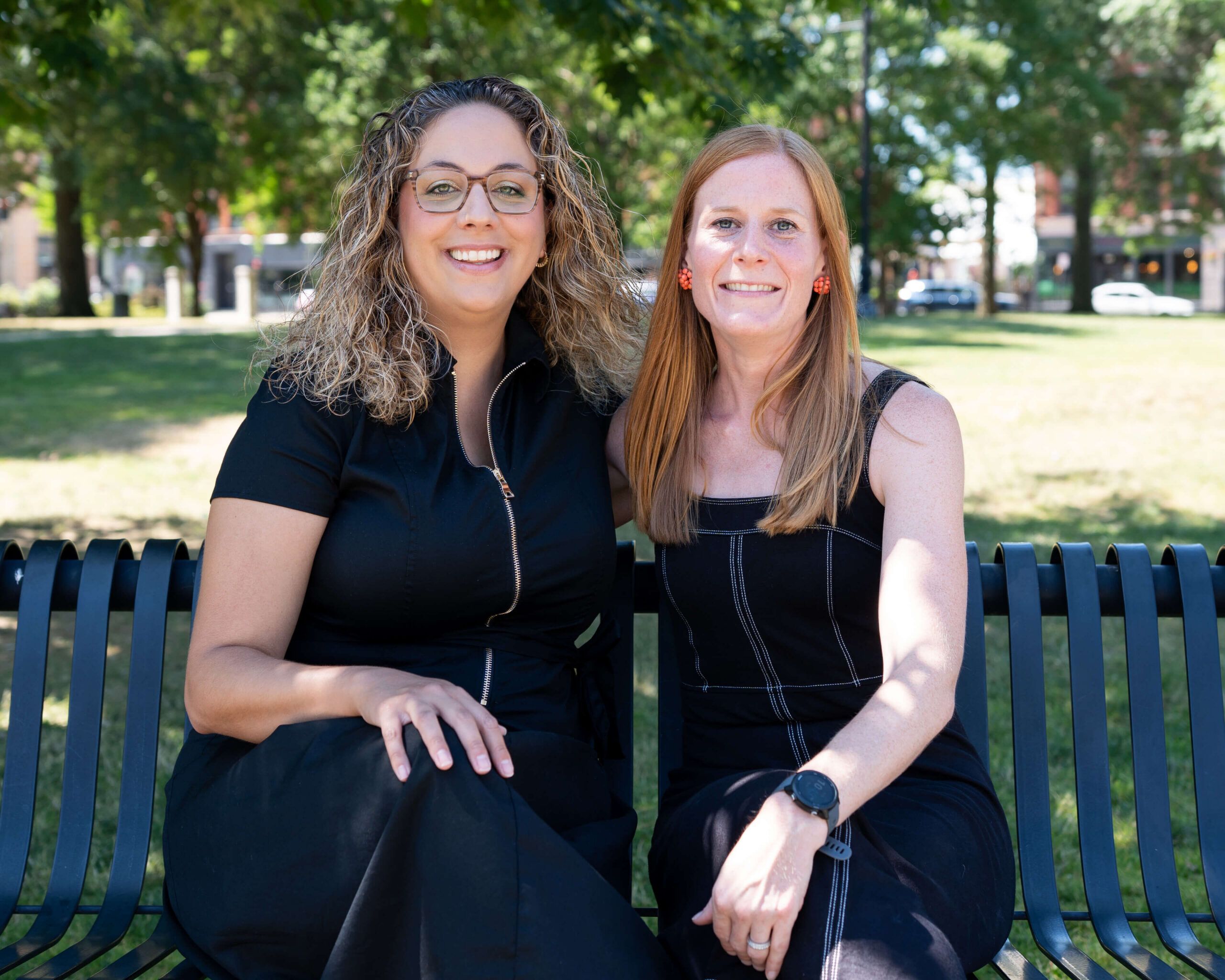 Two women in black dresses sit closely together on a metal bench in a sunny park, smiling at the camera.