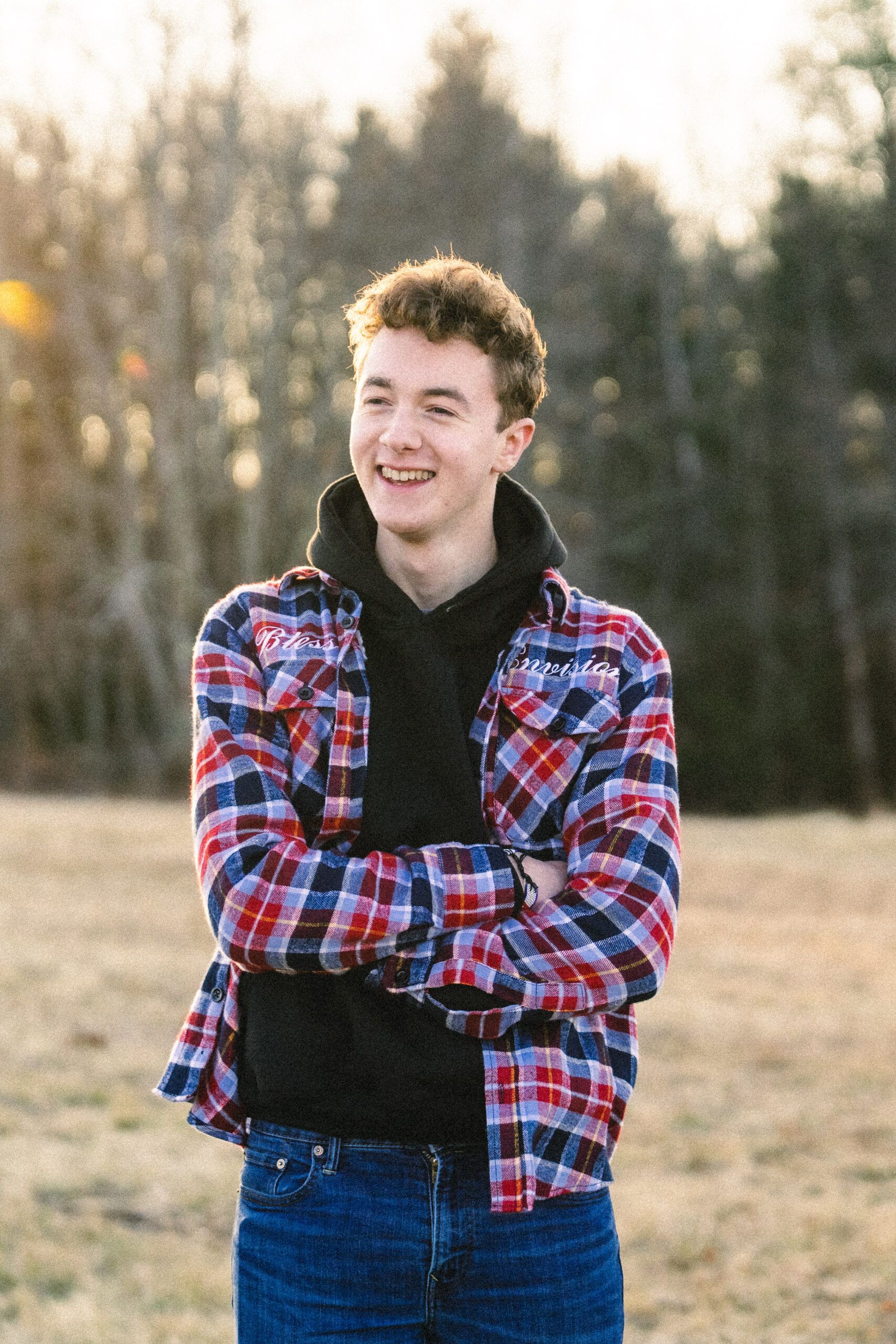 A young person in a plaid shirt and jeans stands outside with arms crossed, smiling, with trees and grass in the background.