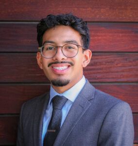 A man with glasses, a mustache, and a goatee smiles while wearing a gray suit, light blue shirt, and dark tie, standing in front of a wooden wall.