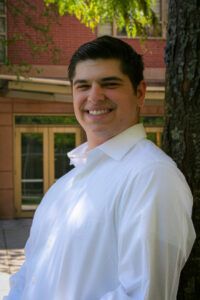 A man in a white dress shirt stands outdoors near a tree, smiling at the camera, with a building and greenery in the background.