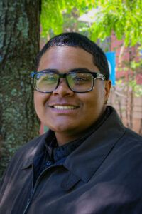 A person in glasses and a dark jacket smiles outdoors near a tree, with green foliage and a blurred building in the background.
