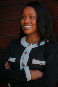 Woman with long dark hair smiles while standing with arms crossed, wearing a black textured jacket with white trim and gold buttons, in front of a dark wooden wall.