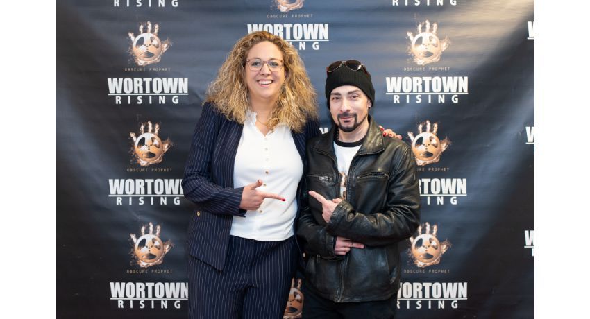 Two people stand in front of a “Wortown Rising” step-and-repeat banner, smiling at the camera and pointing at each other.