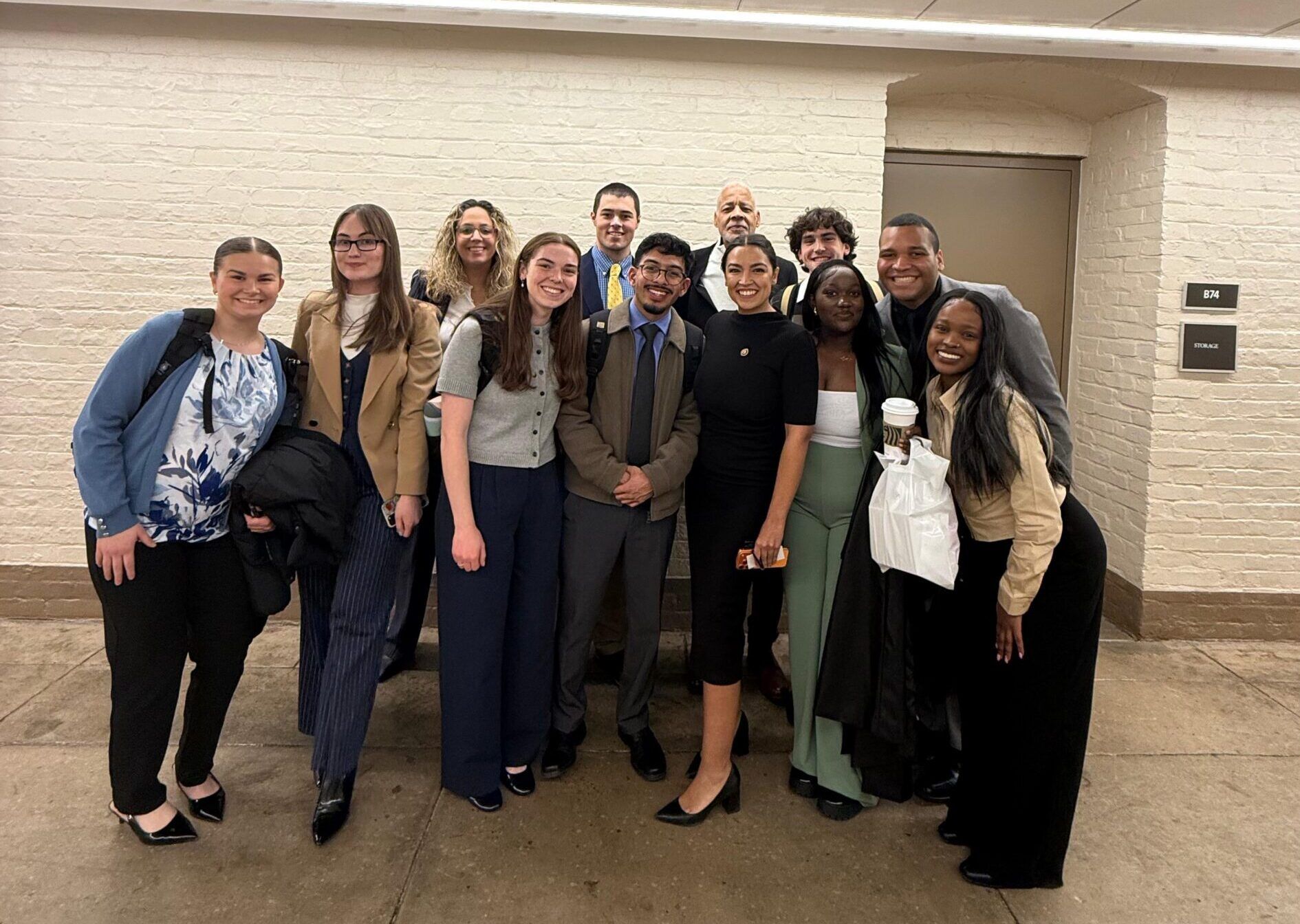 A group of thirteen people stand indoors against a beige brick wall, smiling at the camera.