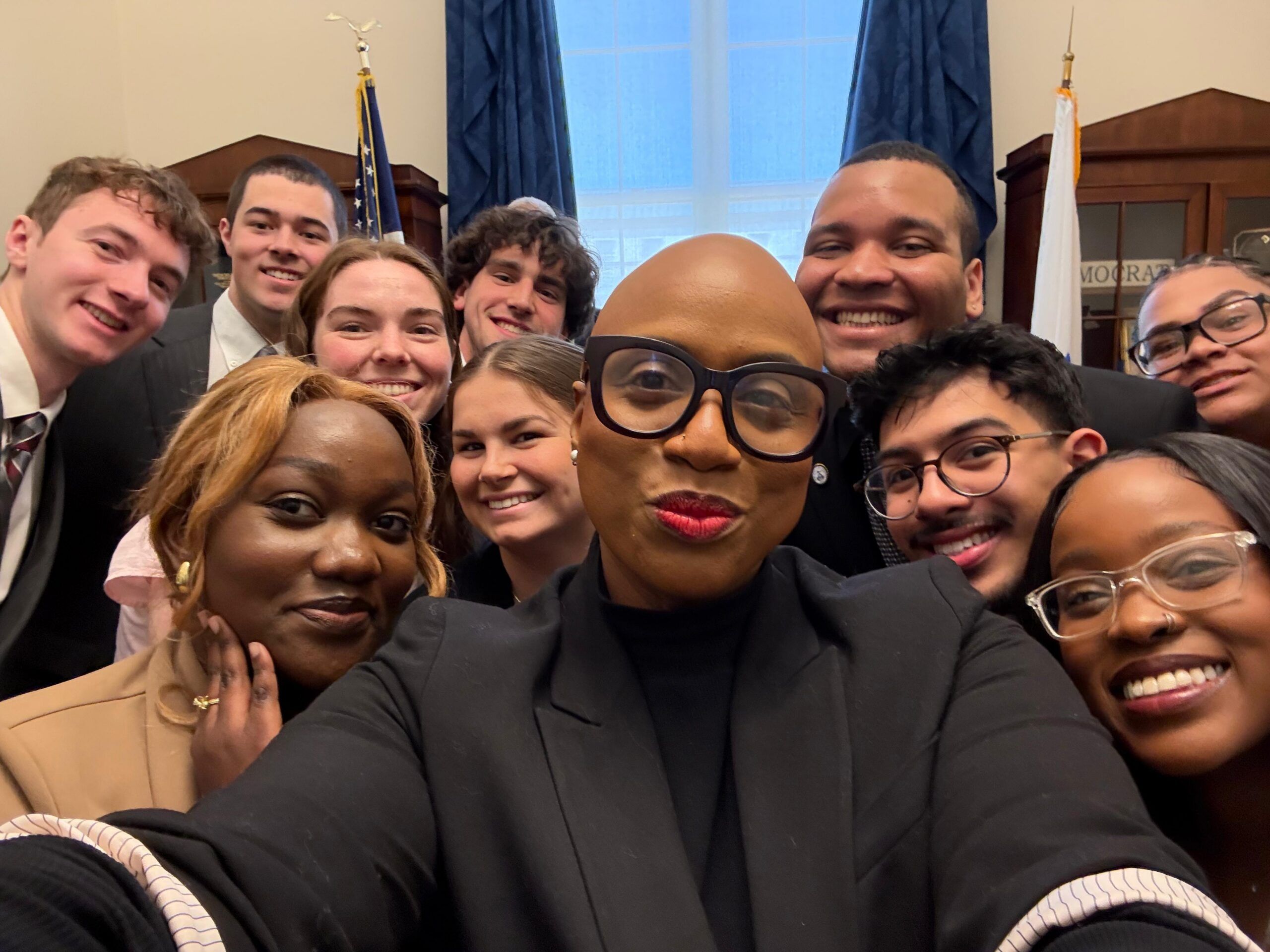 A group of people pose for a selfie indoors, with one person in front holding the camera. Most are smiling, and there are flags and blue curtains in the background.