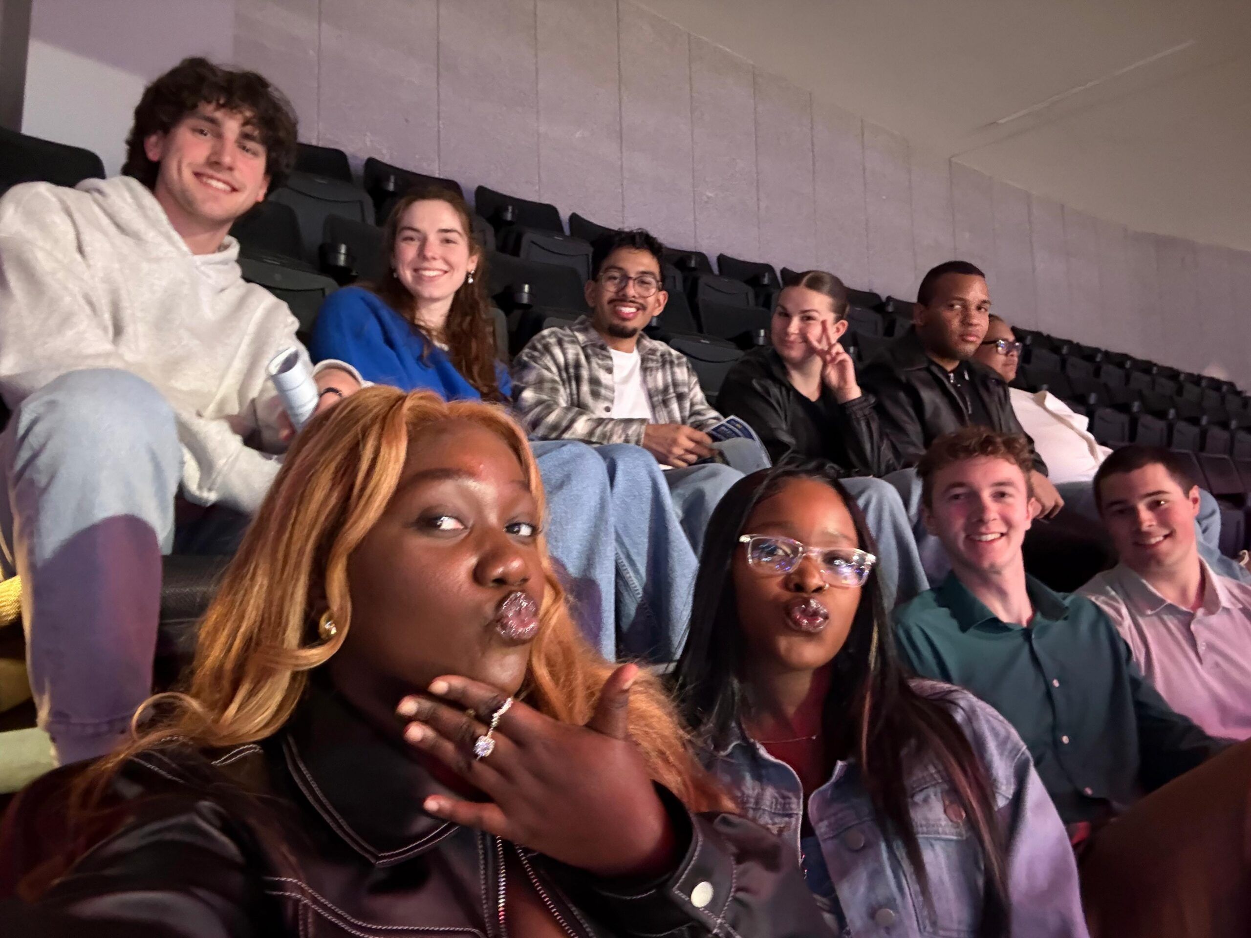 A group of ten young adults sits in stadium-style seats, with two in front taking a selfie. Most are smiling and dressed casually.