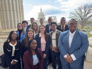 A group of people posing together on a balcony with the US Capitol building visible in the background on a partly cloudy day.