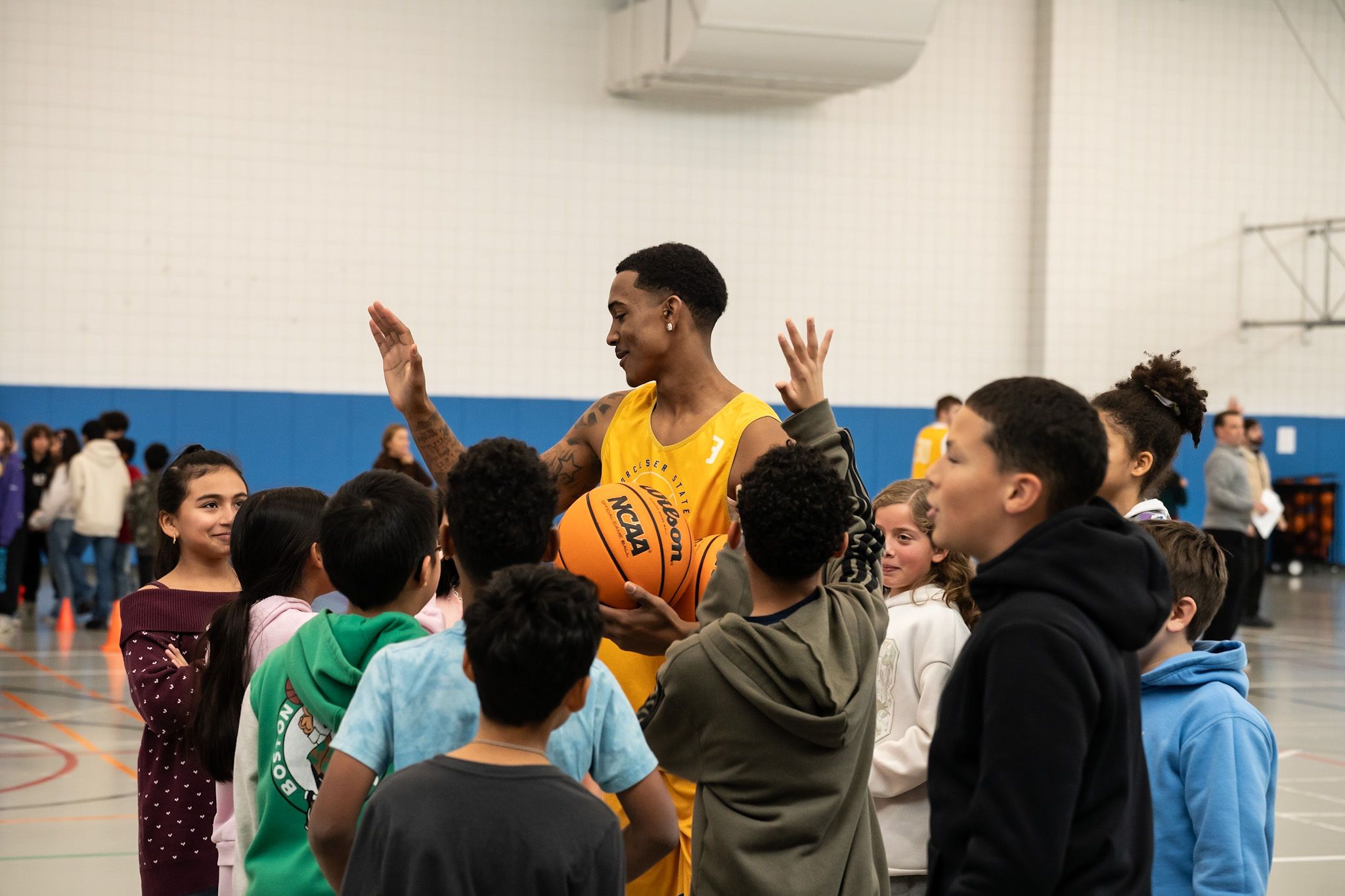 A basketball player in a yellow jersey holding a ball smiles and high-fives a group of children in a gymnasium.