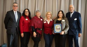 Six people stand in a row indoors, five women and one man, with one woman holding a framed award; all are dressed in business or business casual attire.