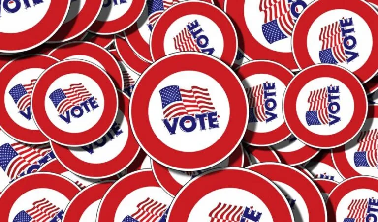 A pile of round buttons with red borders displaying an American flag and the word “VOTE” in blue capital letters.
