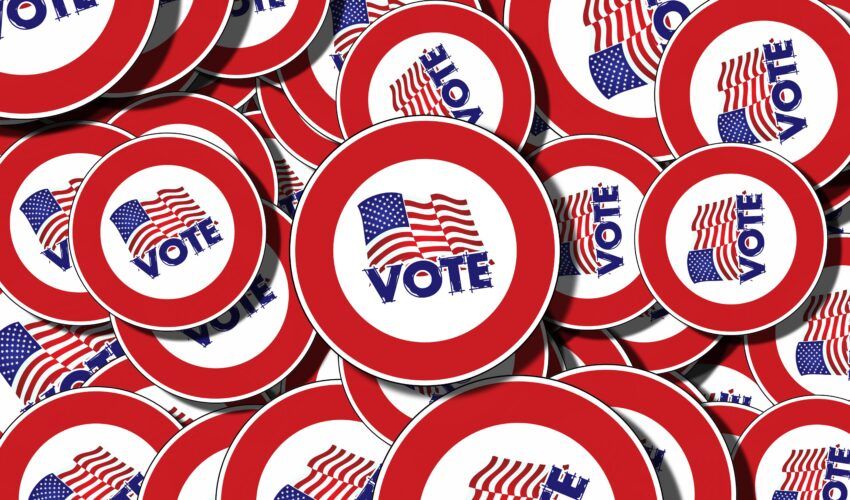 A pile of round buttons with a red border, featuring an American flag and the word “VOTE” in blue capital letters in the center.