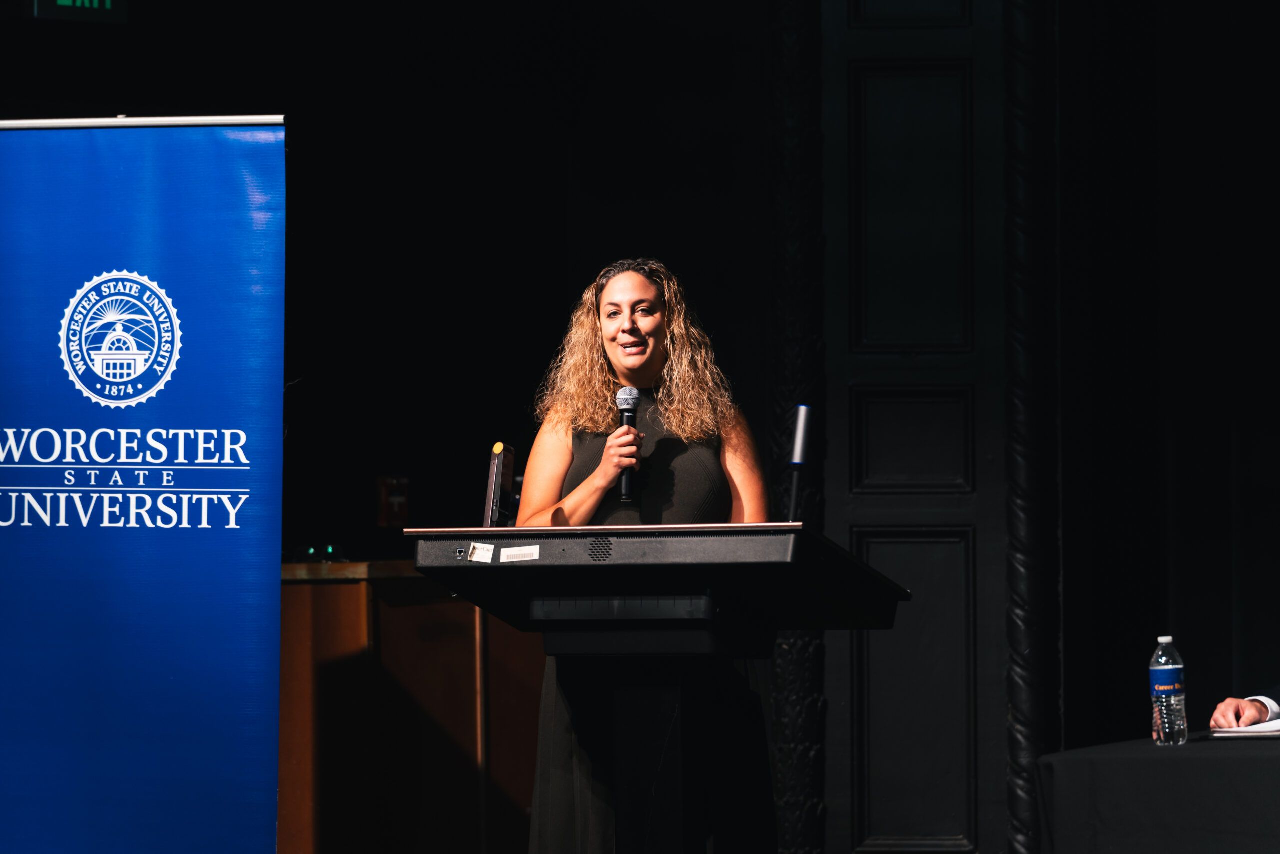 A woman stands at a podium holding a microphone, with a Worcester State University banner to her left.
