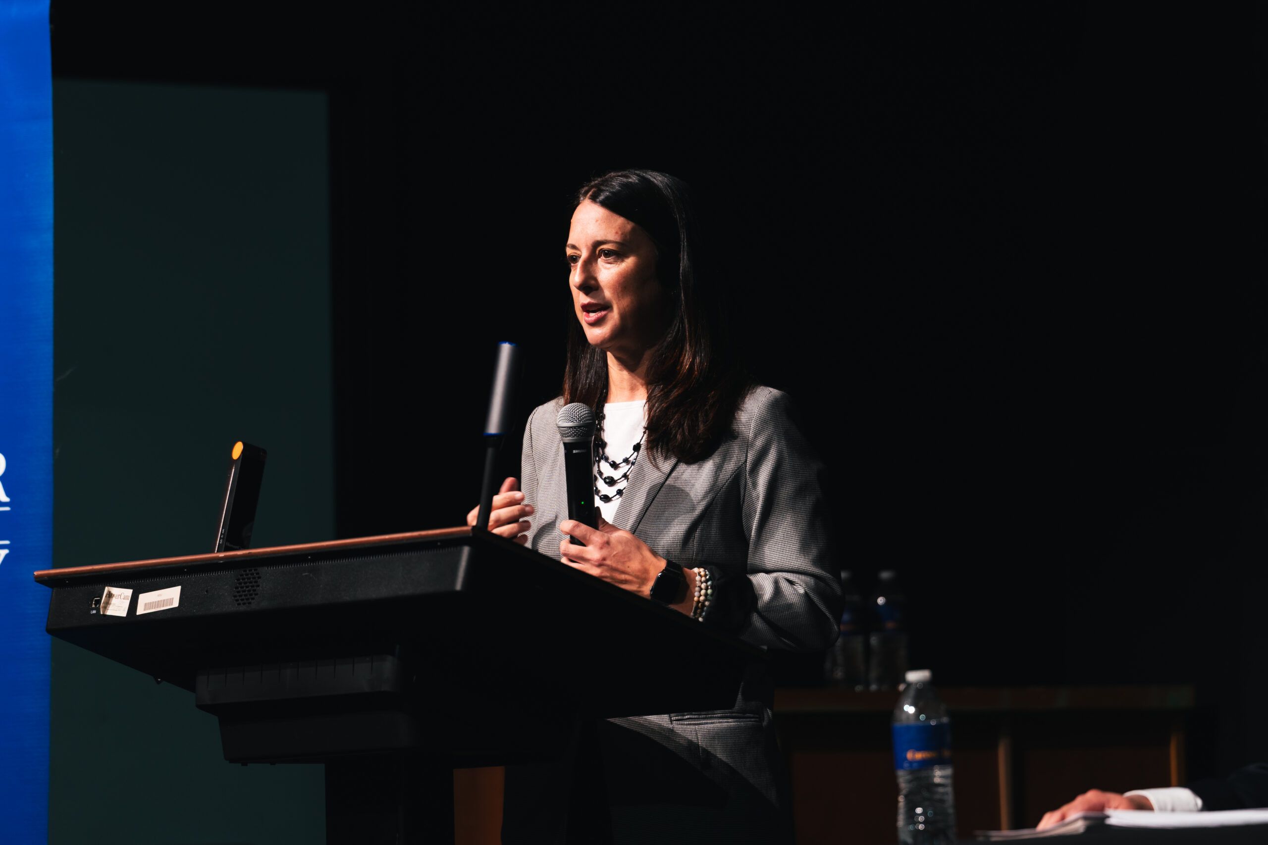 A woman in a grey blazer stands at a podium speaking into a microphone.