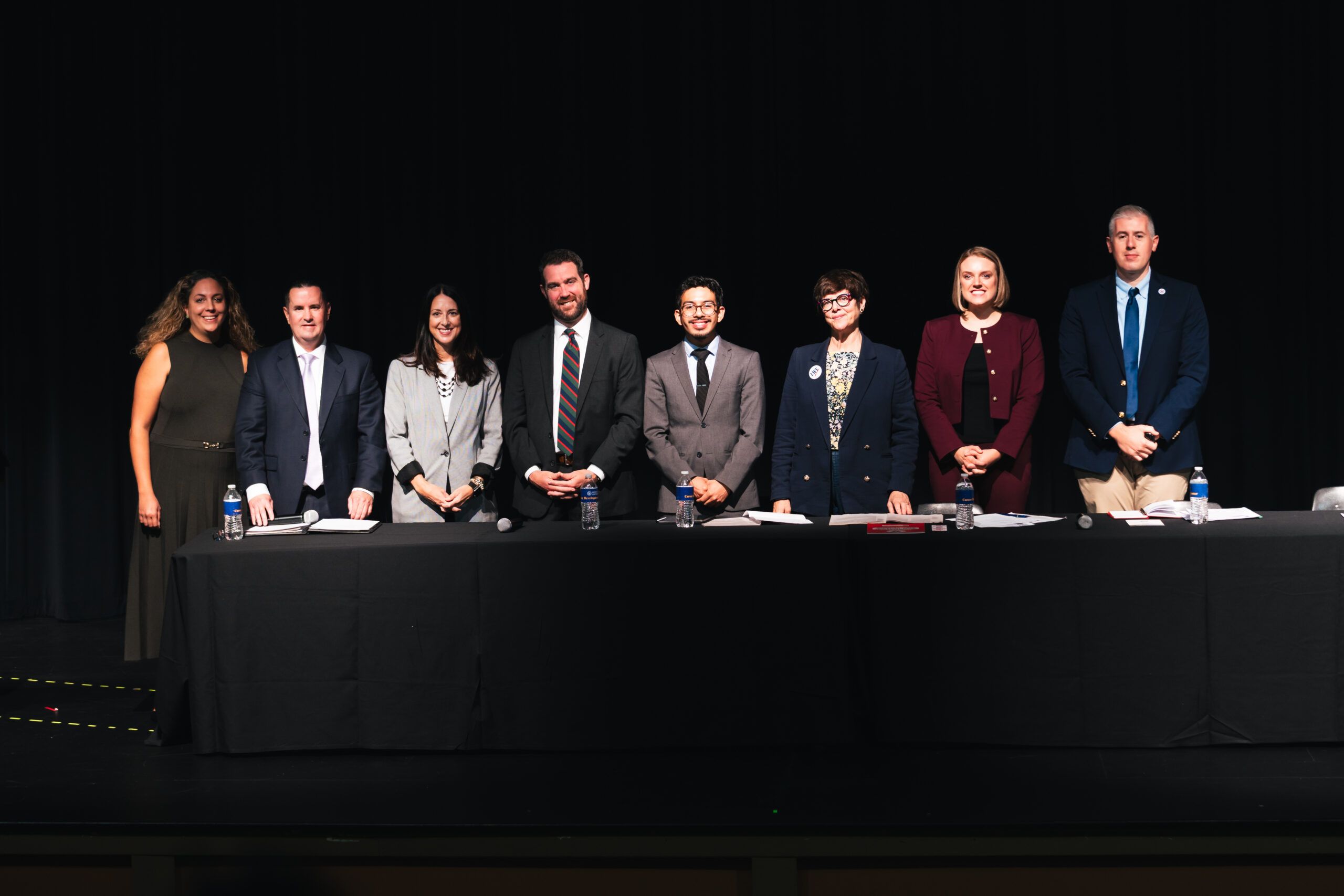 Seven people stand behind a long table on a stage against a black background.
