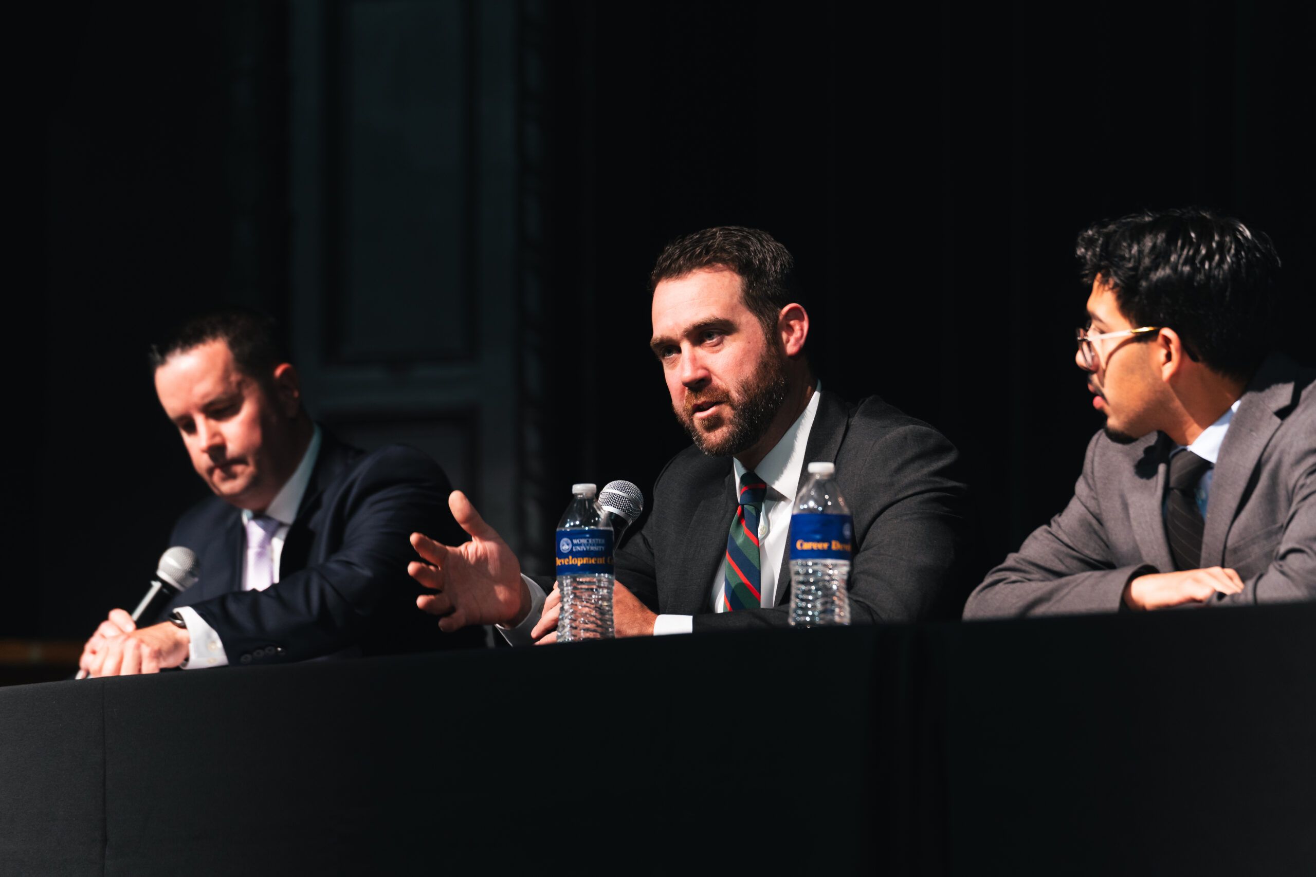 Three men in suits sitting at a table on a stage during a panel discussion.