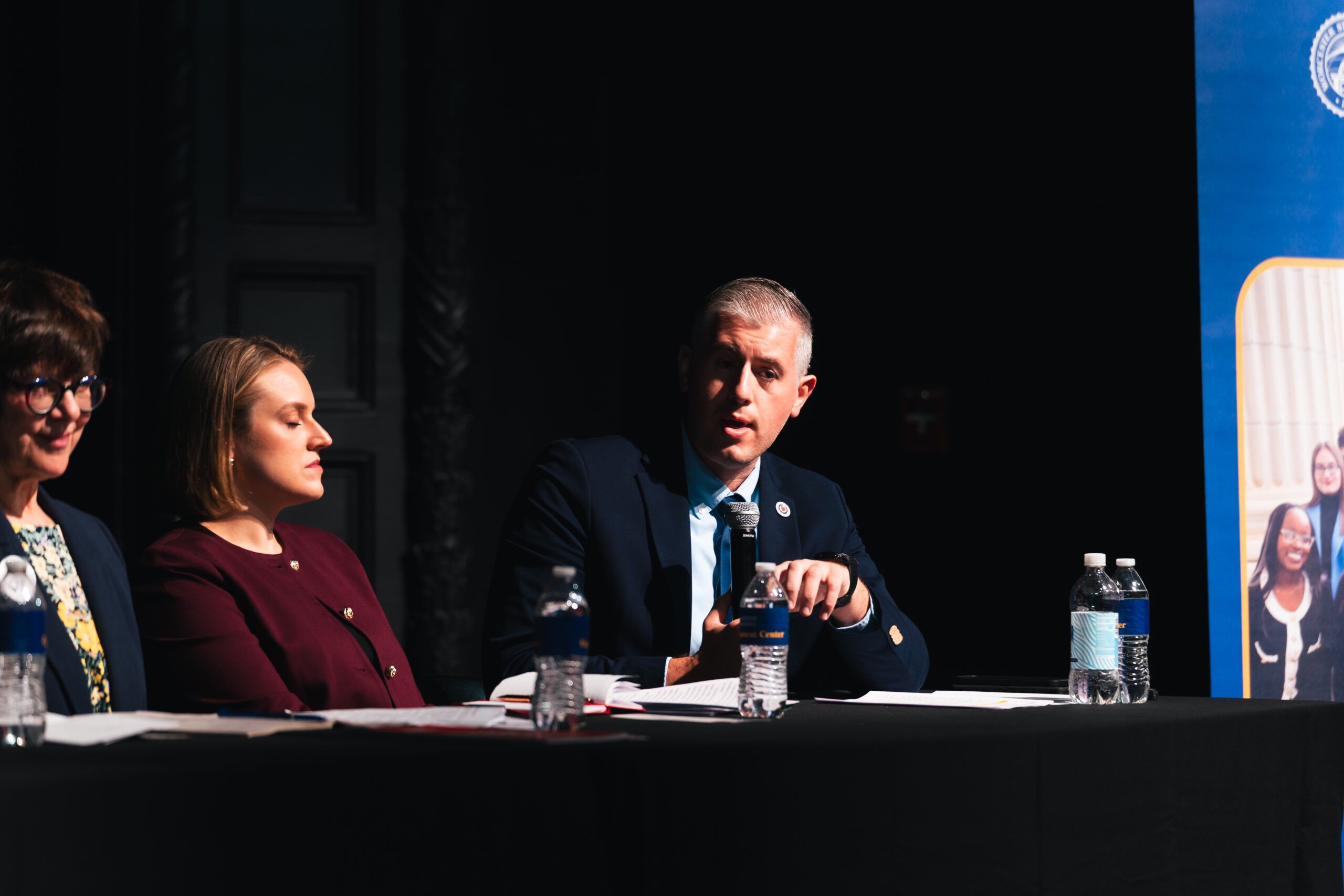 A panel of three people seated at a table, with the man in the center speaking into a microphone.