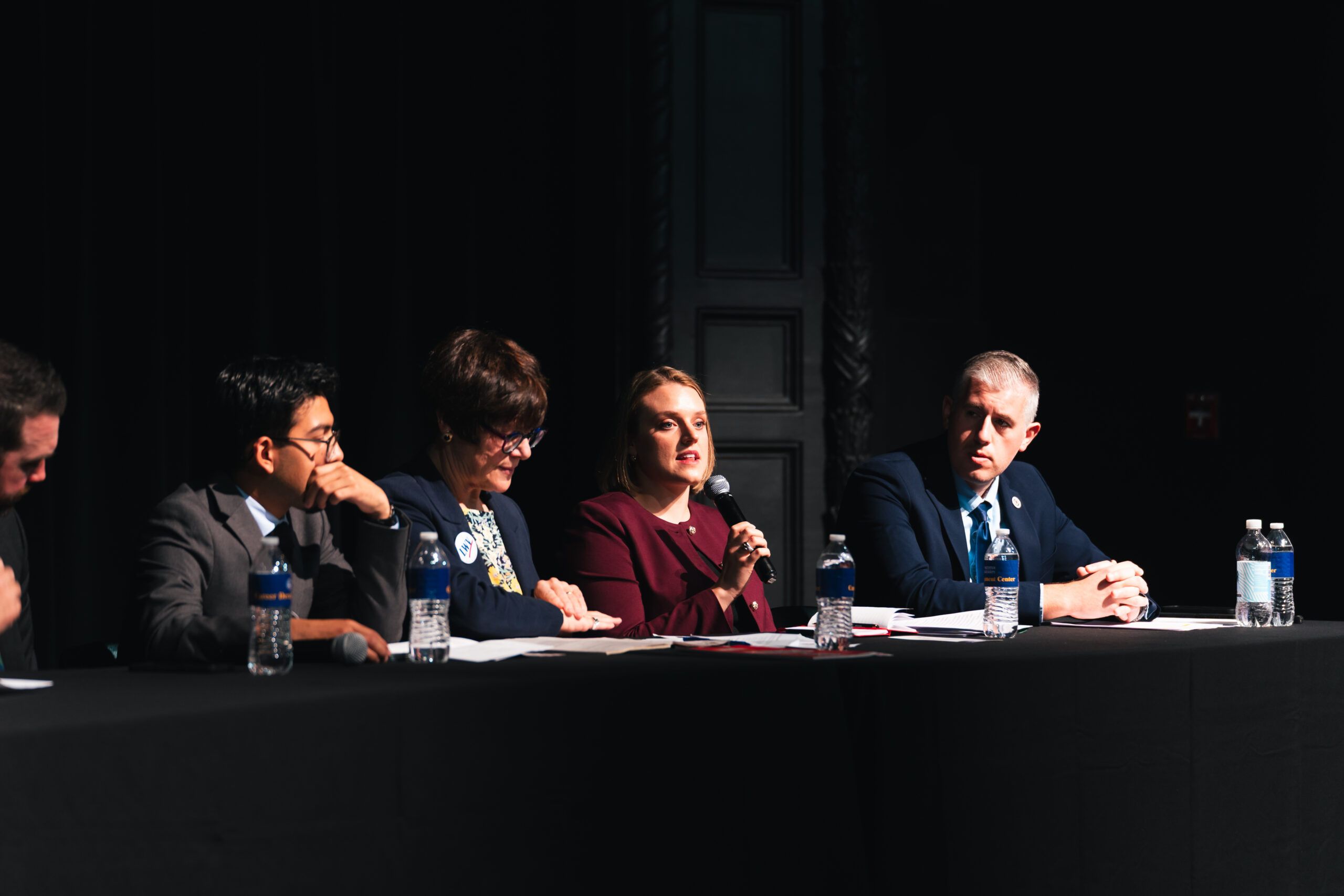 A panel of four people seated at a table on a dark stage, with one woman speaking into a microphone.