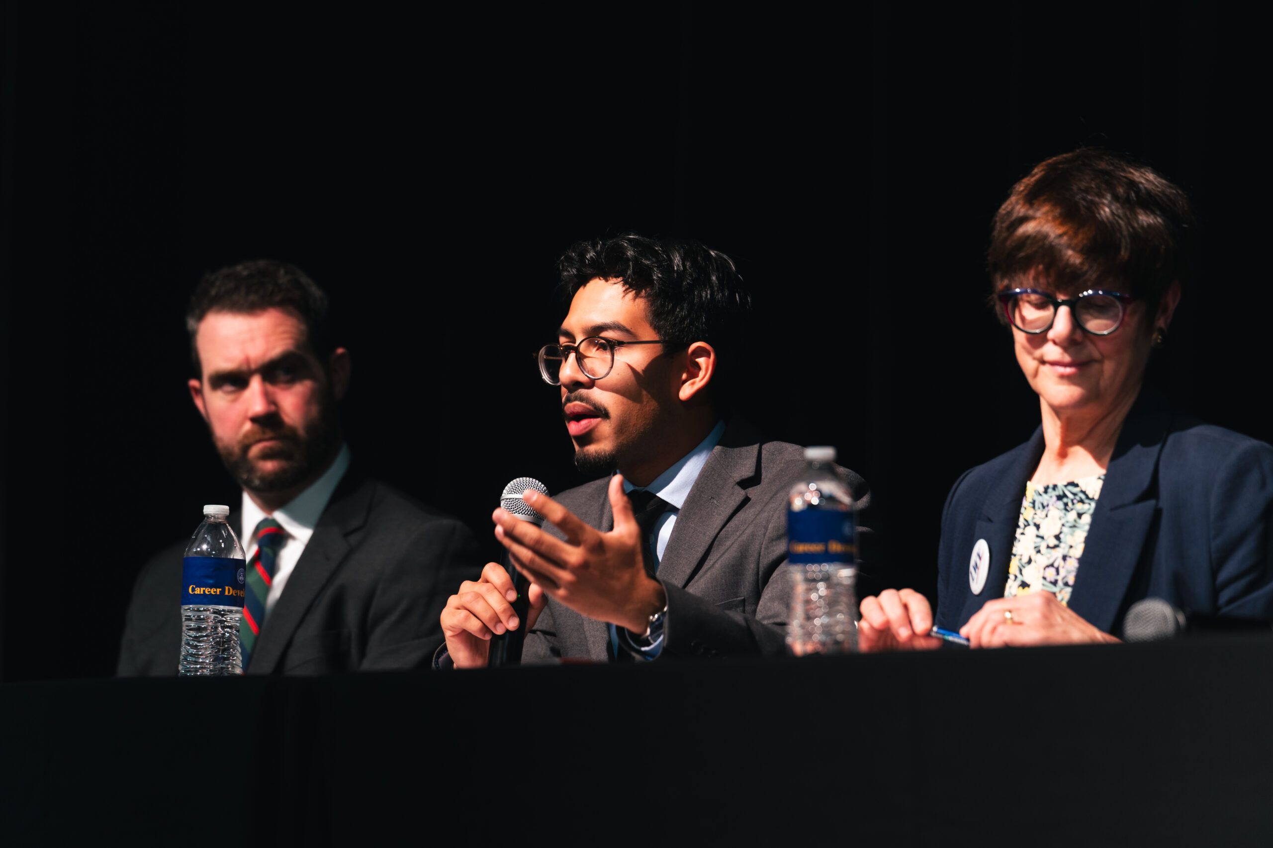 A panel of three people sit at a table on a stage, with the center person speaking into a microphone.