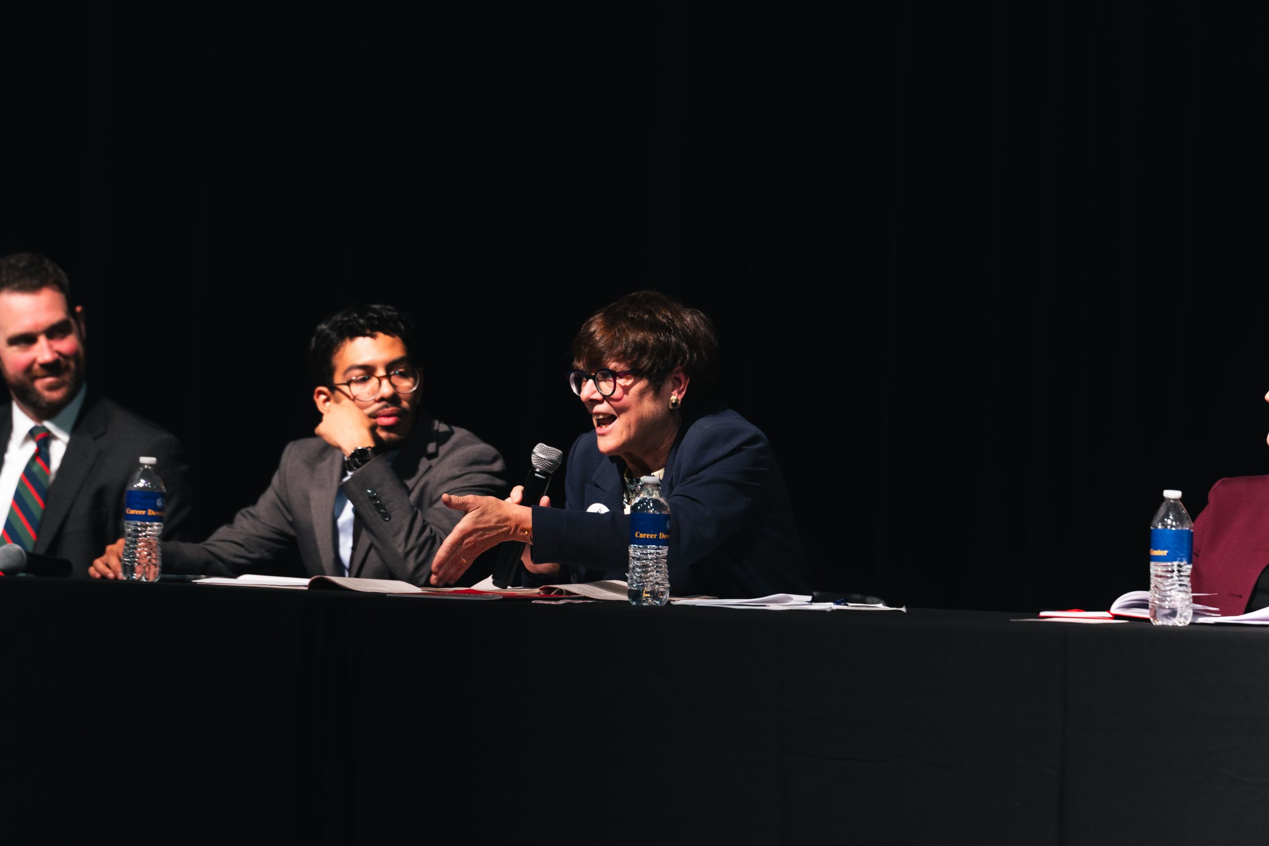 A woman speaks into a microphone at a panel discussion with two men seated beside her at a long table.