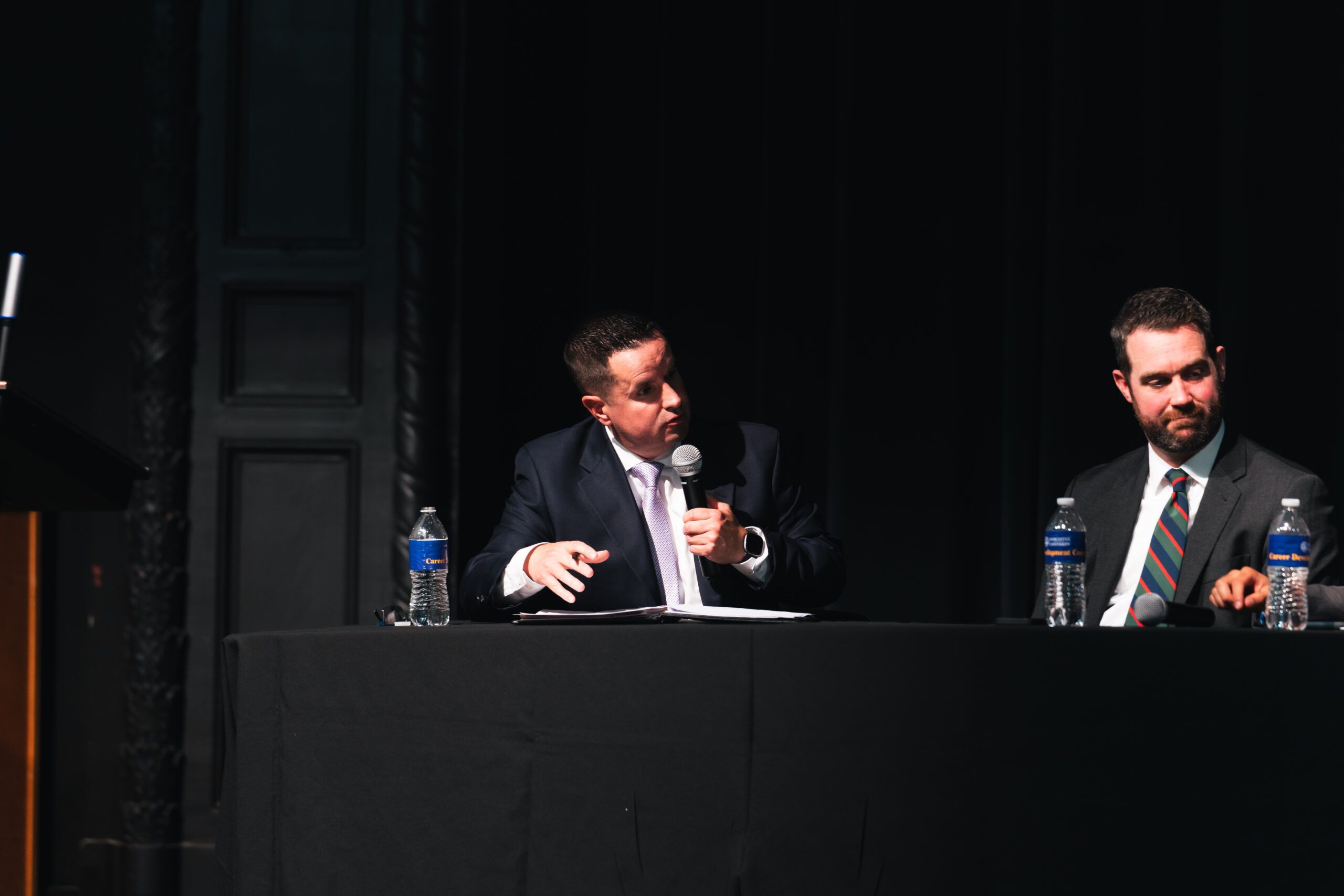 Two men in suits sit at a table on a stage, with one holding a microphone and speaking.
