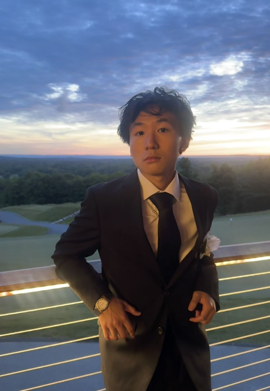 A young man in a black suit and tie standing on a balcony overlooking a golf course at sunset.
