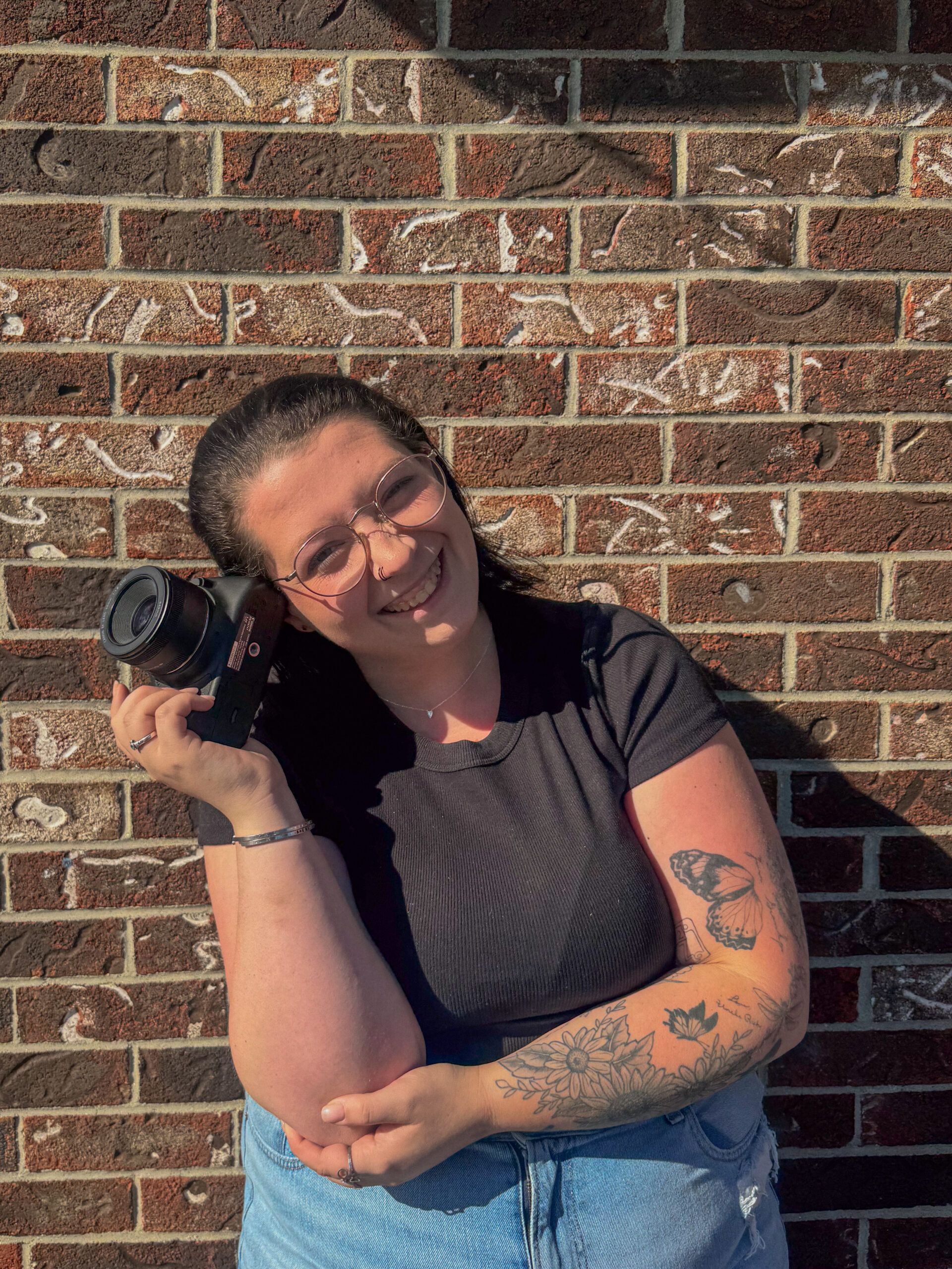 A smiling woman wearing glasses and holding a camera against her ear, posing in front of a brick wall.