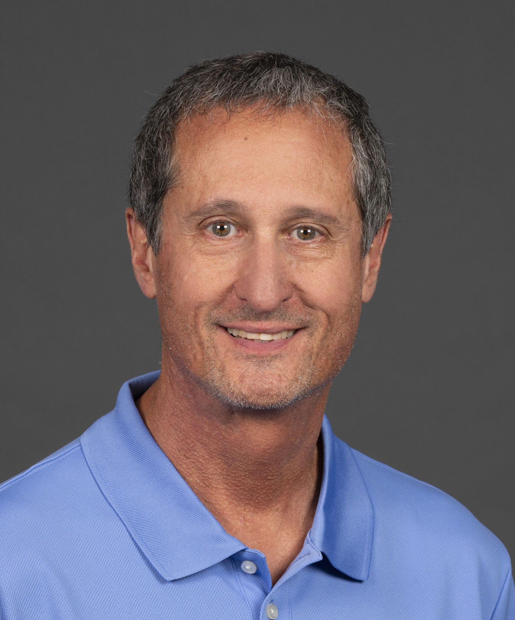 A man in a blue polo shirt smiles against a plain gray background.