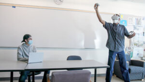 Student wearing VR goggles, while another engages on laptop nearby